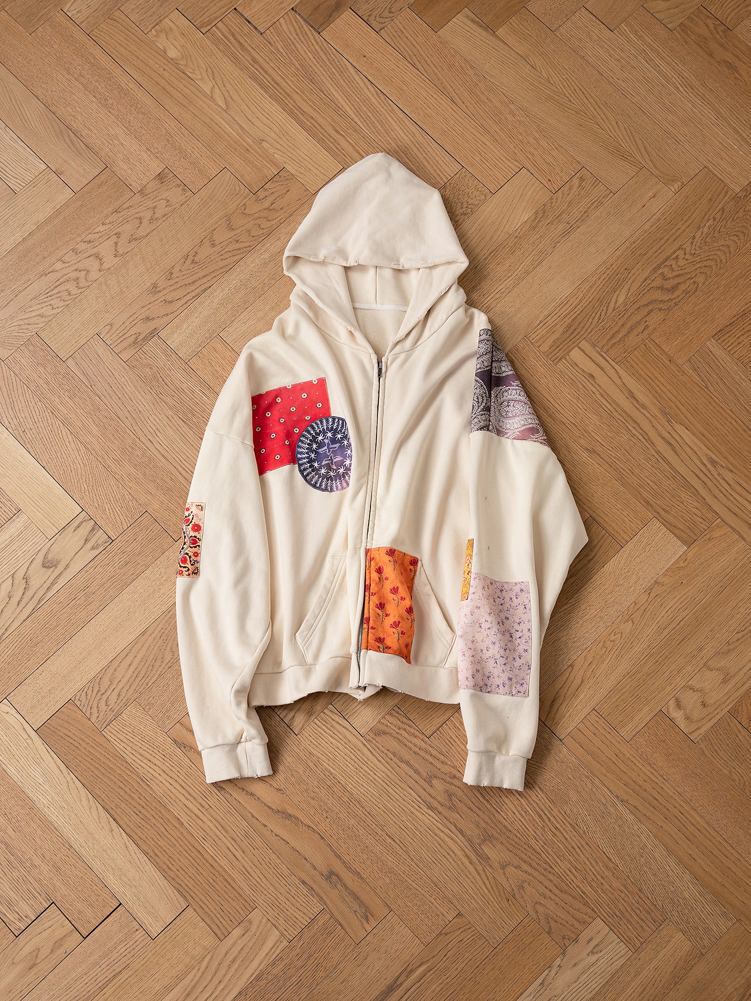 The Tapestry Patchwork Zip Up Hoodie by FOUND showcases vibrant South Asian tapestry patterns and is elegantly displayed on a wooden herringbone floor.