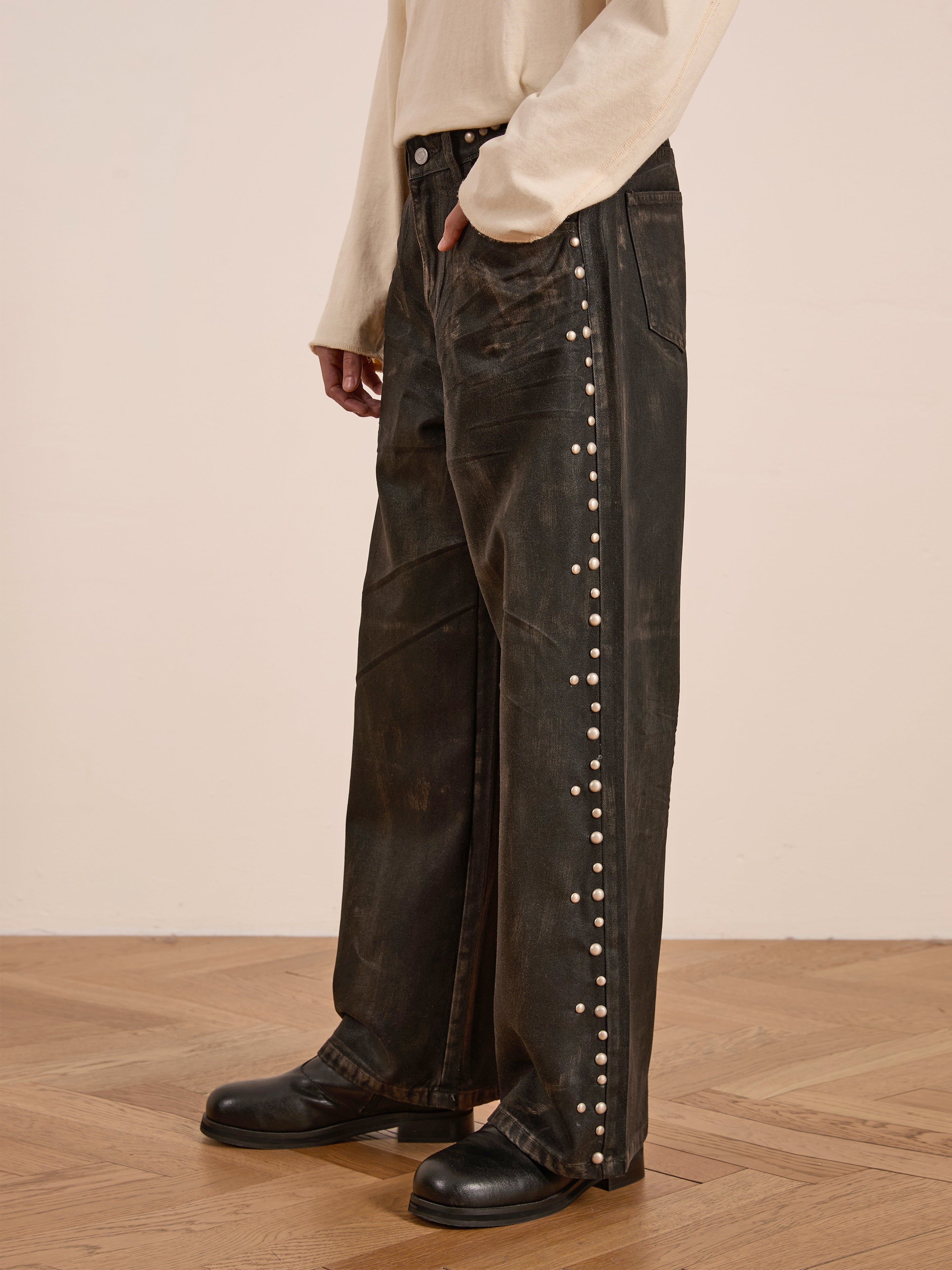 A person stands on a wooden floor wearing FOUND's Waxed Studded Jeans with pearl-like accents down the sides, paired with a cream long-sleeve shirt and black shoes. The ensemble evokes vintage craftsmanship with its intricate details.