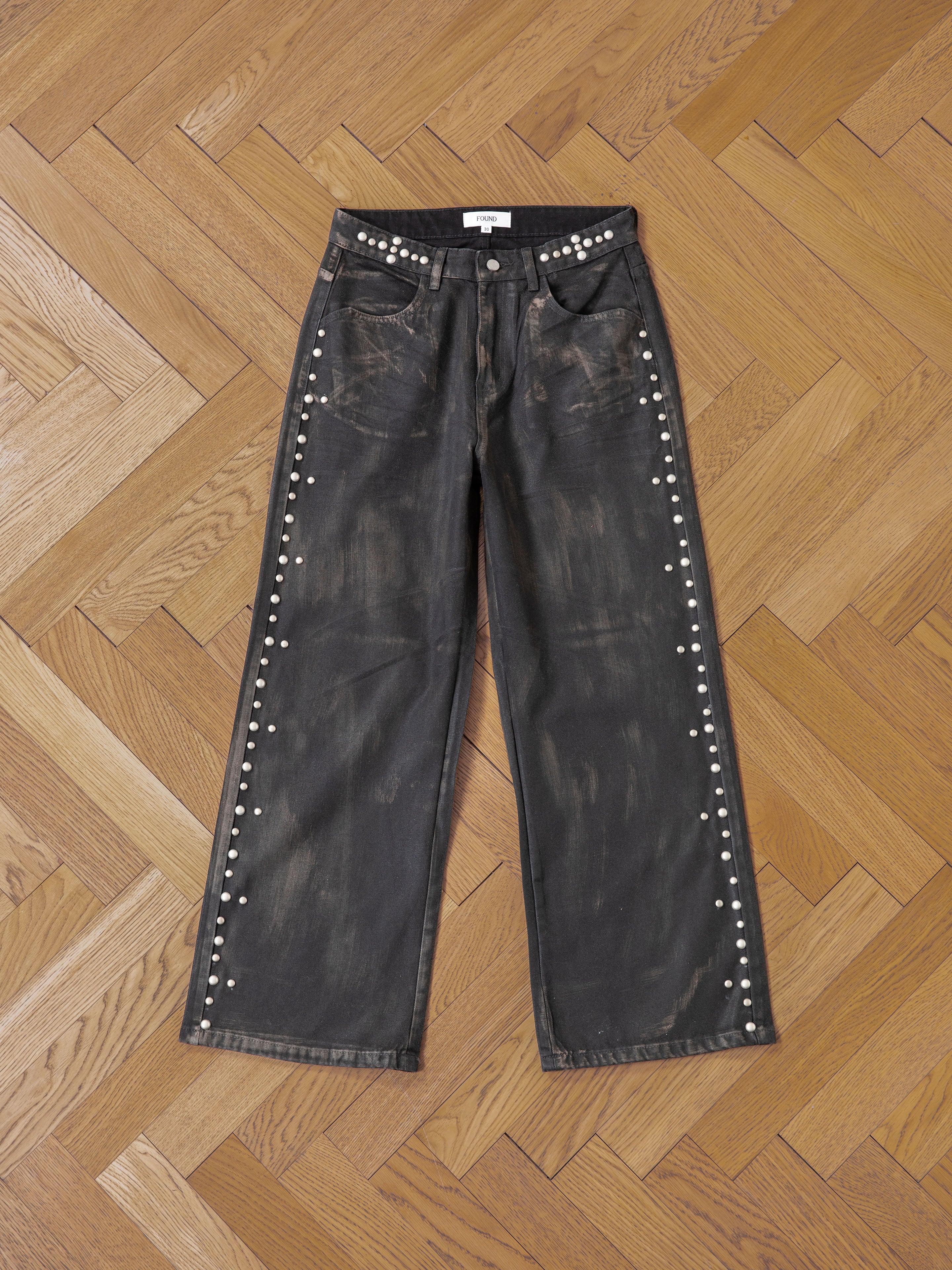 The Waxed Studded Jeans by FOUND feature a vintage craftsmanship vibe with decorative white studs along the sides. This unisex, wide-leg, relaxed-fit piece is both stylish and comfortable for any occasion, displayed on a wooden herringbone parquet floor.