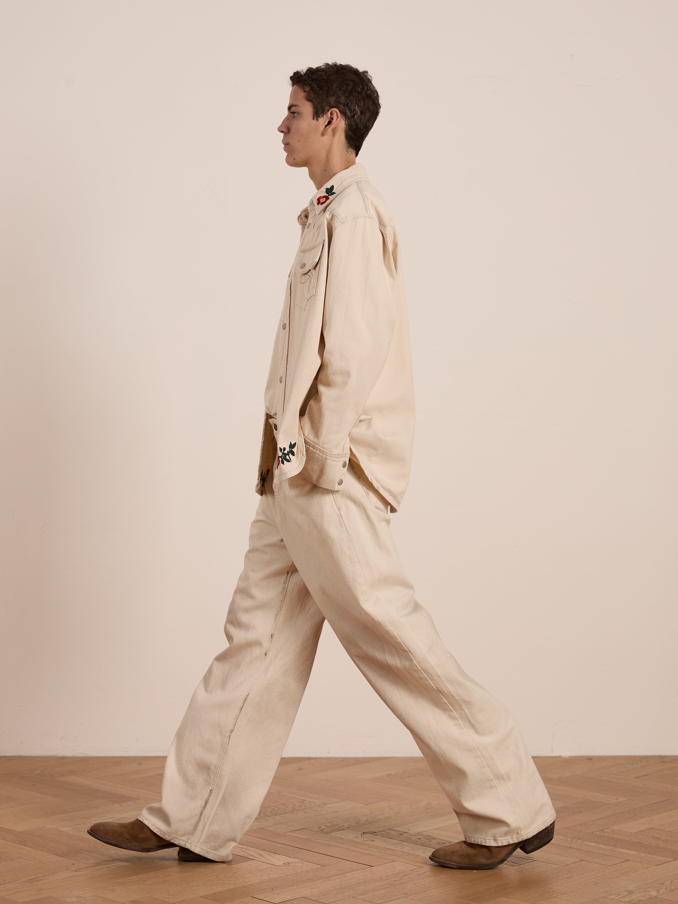 A person in a Western Floral Denim Shirt by FOUND and brown shoes walks sideways on a wooden floor, the attire subtly complemented by its floral chainstitch embroidery against a plain wall.