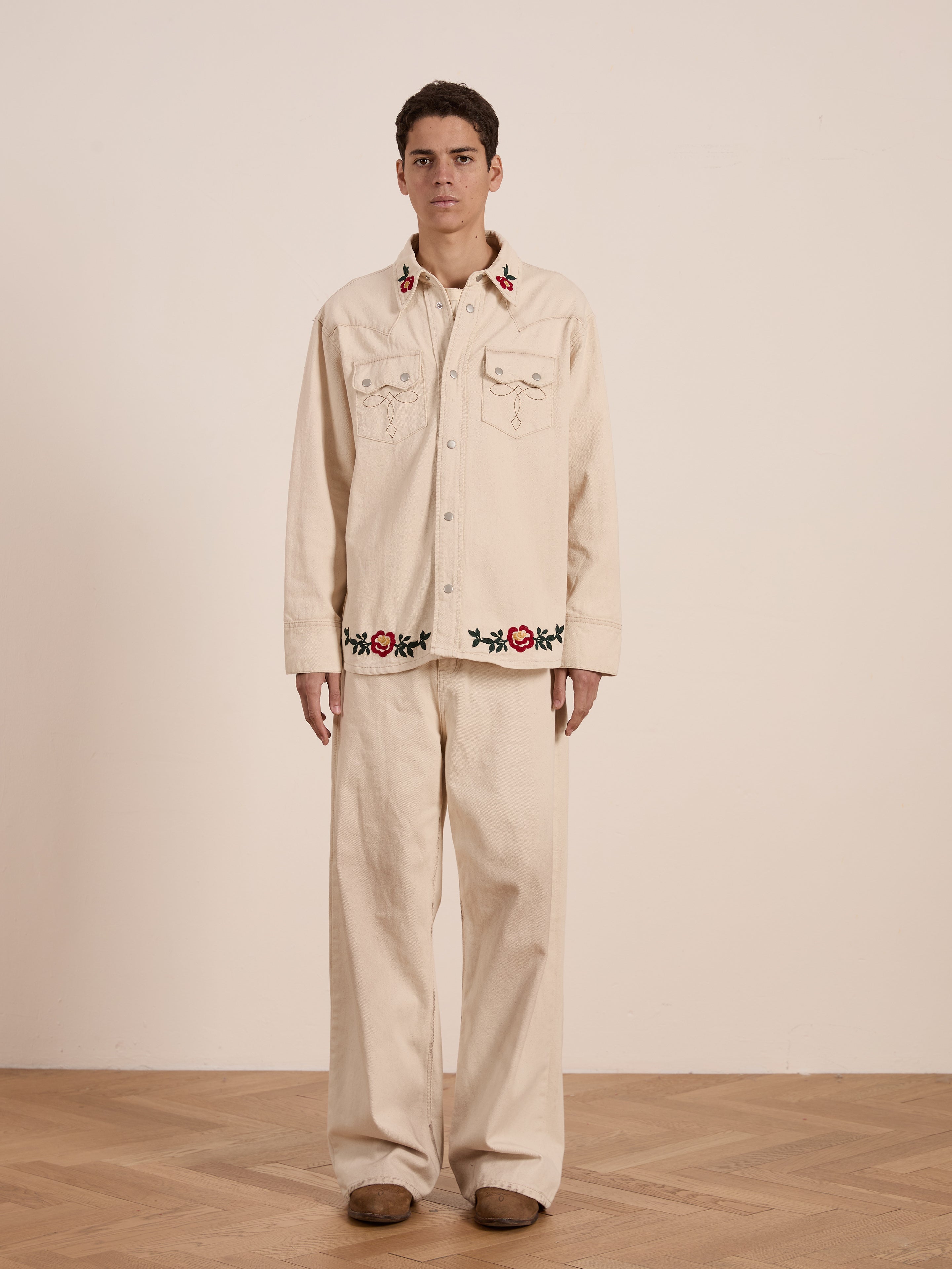 A person stands in the room wearing a beige outfit featuring intricate floral chainstitch embroidery, complemented by brown shoes and the Western Floral Denim Shirt by FOUND.