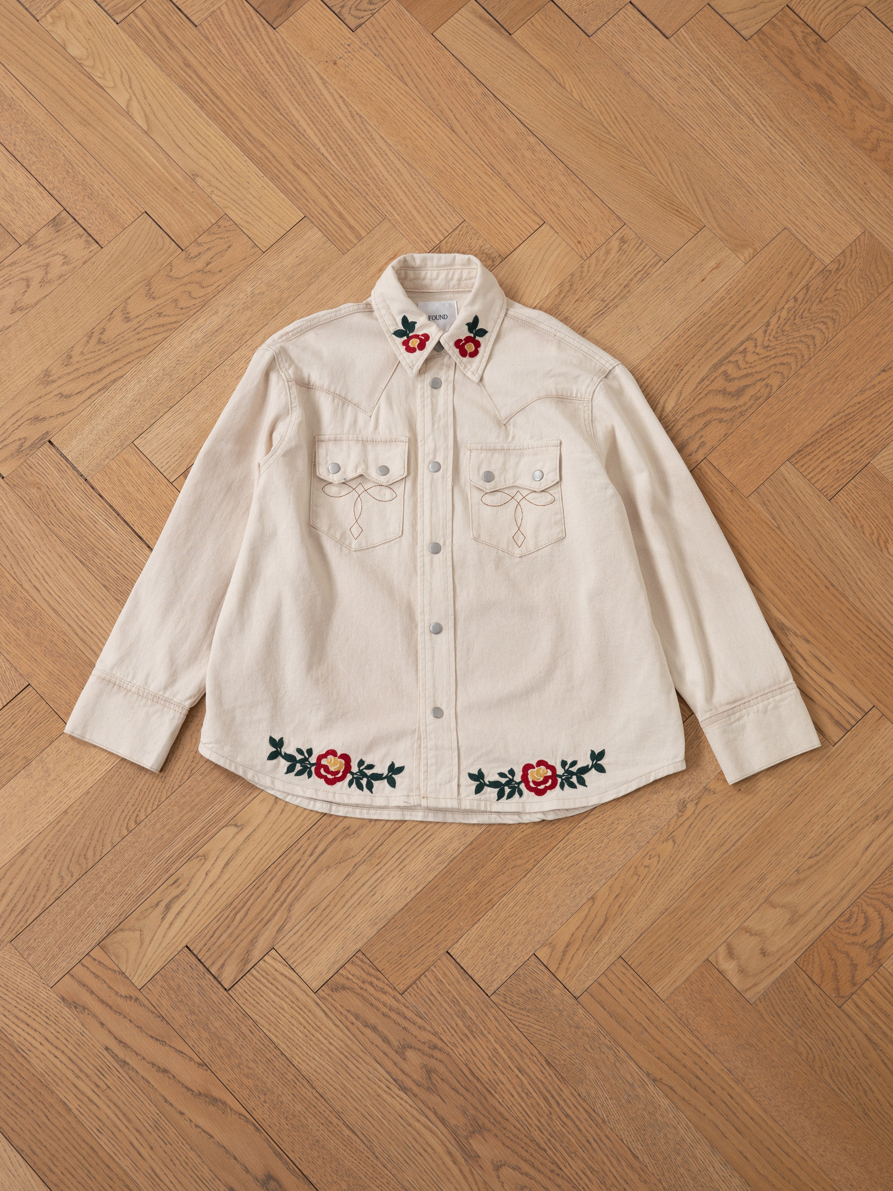 Laid flat on a wooden herringbone-pattern floor, this beige button-up shirt from FOUND features intricate floral chainstitch embroidery on the collar and bottom, subtly nodding to the Western Floral Denim Shirt style.