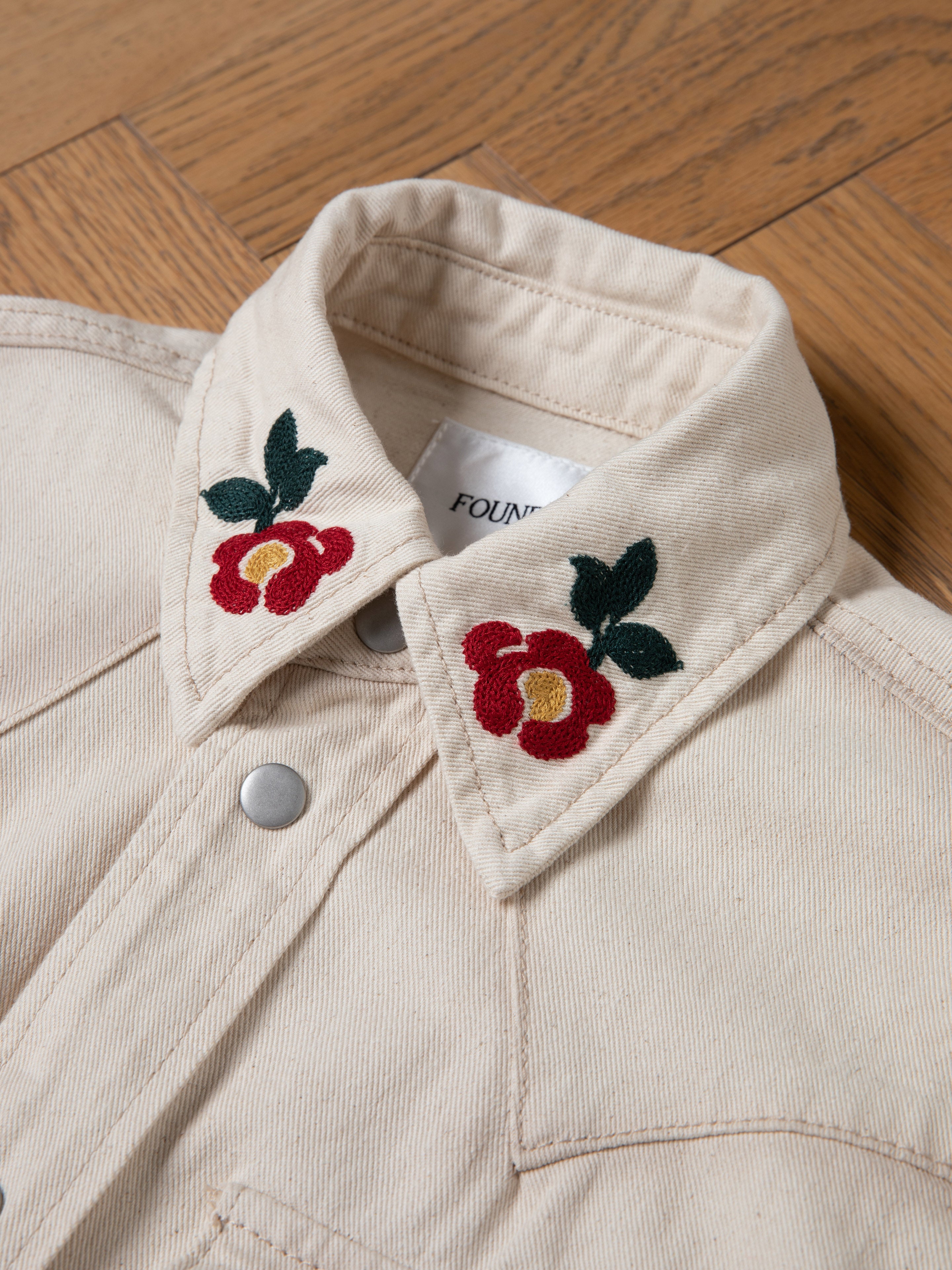 FOUND's Western Floral Denim Shirt in soft-washed cream features red floral chainstitch embroidery on the collar and metal snap buttons, elegantly showcased on a wooden surface.