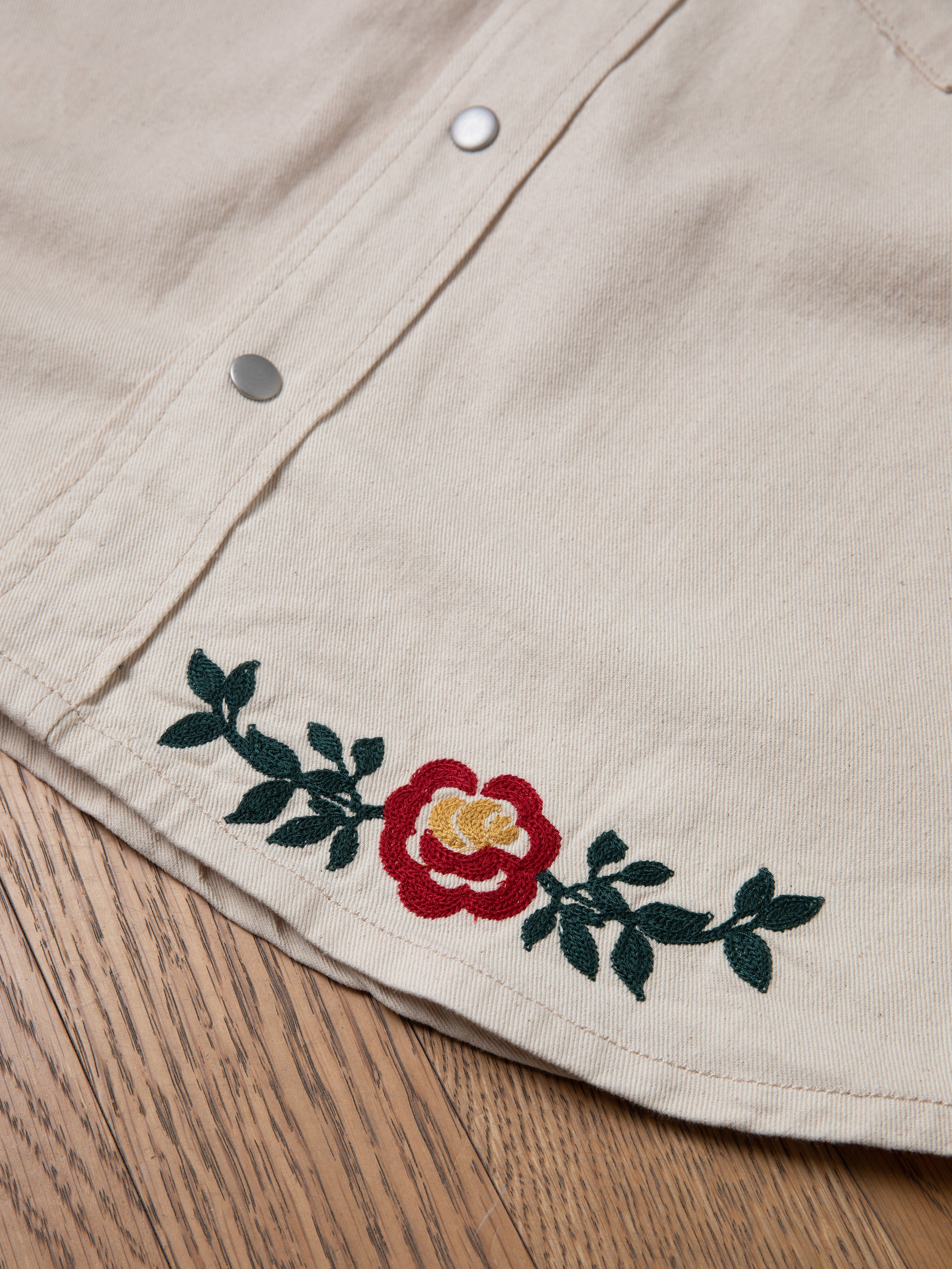 Beige fabric with red and yellow floral chainstitch embroidery and green leaves, accented by metal button snaps, beautifully displays the charm of FOUND's Western Floral Denim Shirt on a wooden surface.