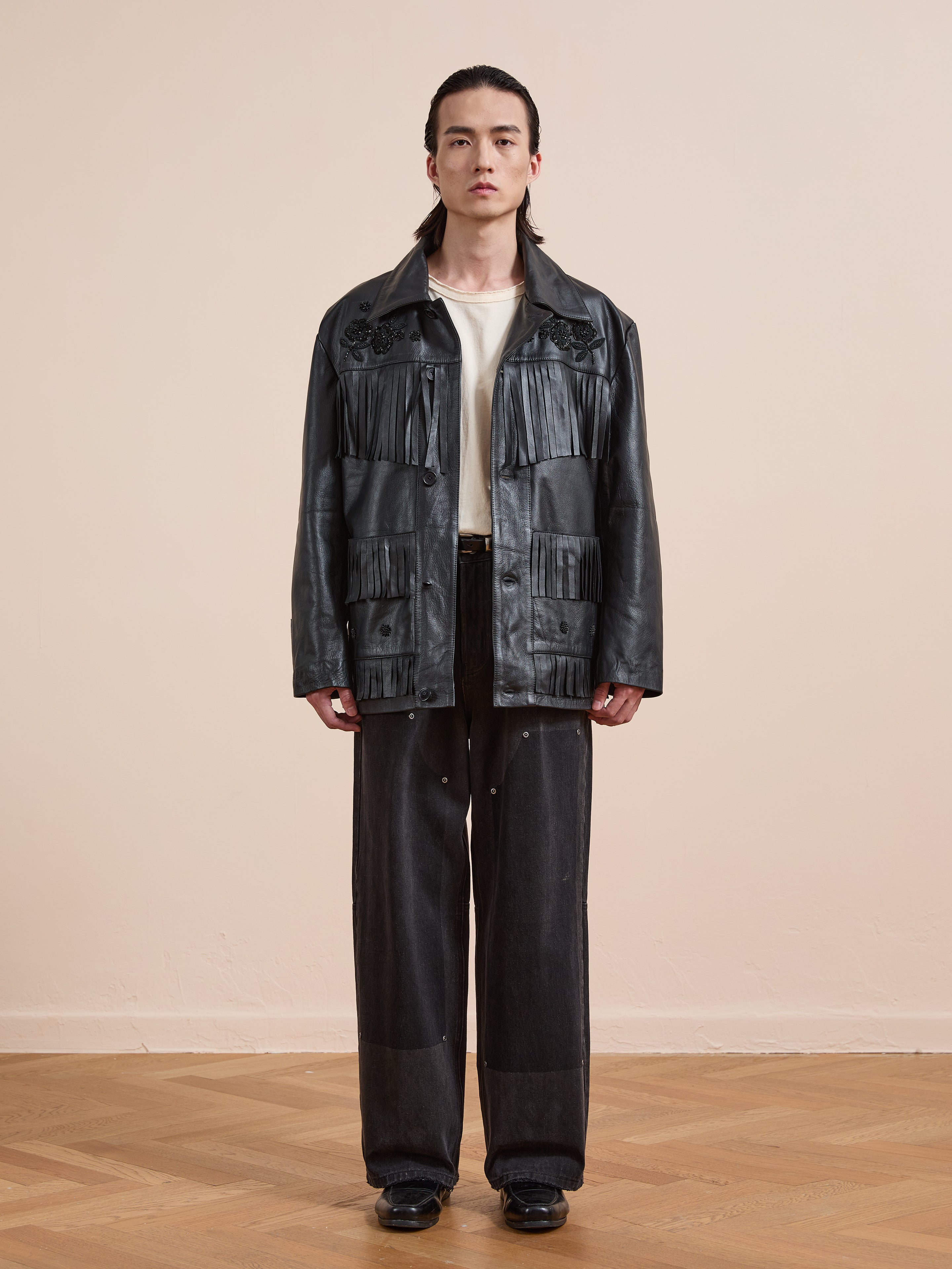 A person stands indoors against a plain beige wall, wearing the FOUND Hand Embroidered Leather Fringe Jacket, a white shirt, loose black pants, and black shoes.