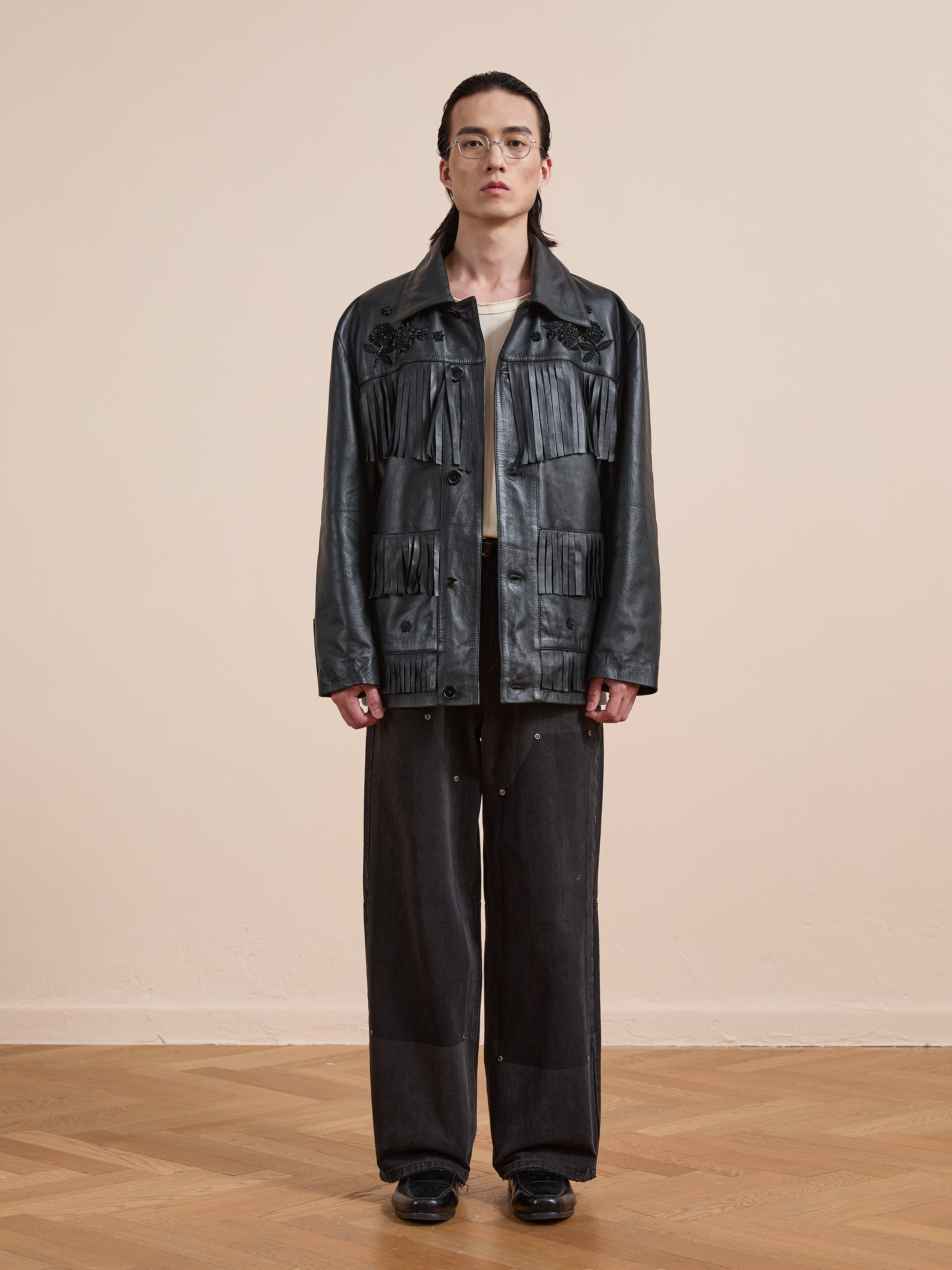 Indoors on a wooden floor, a person wearing glasses styles the FOUND Hand Embroidered Leather Fringe Jacket with wide-leg dark pants and black shoes, standing against a beige background.