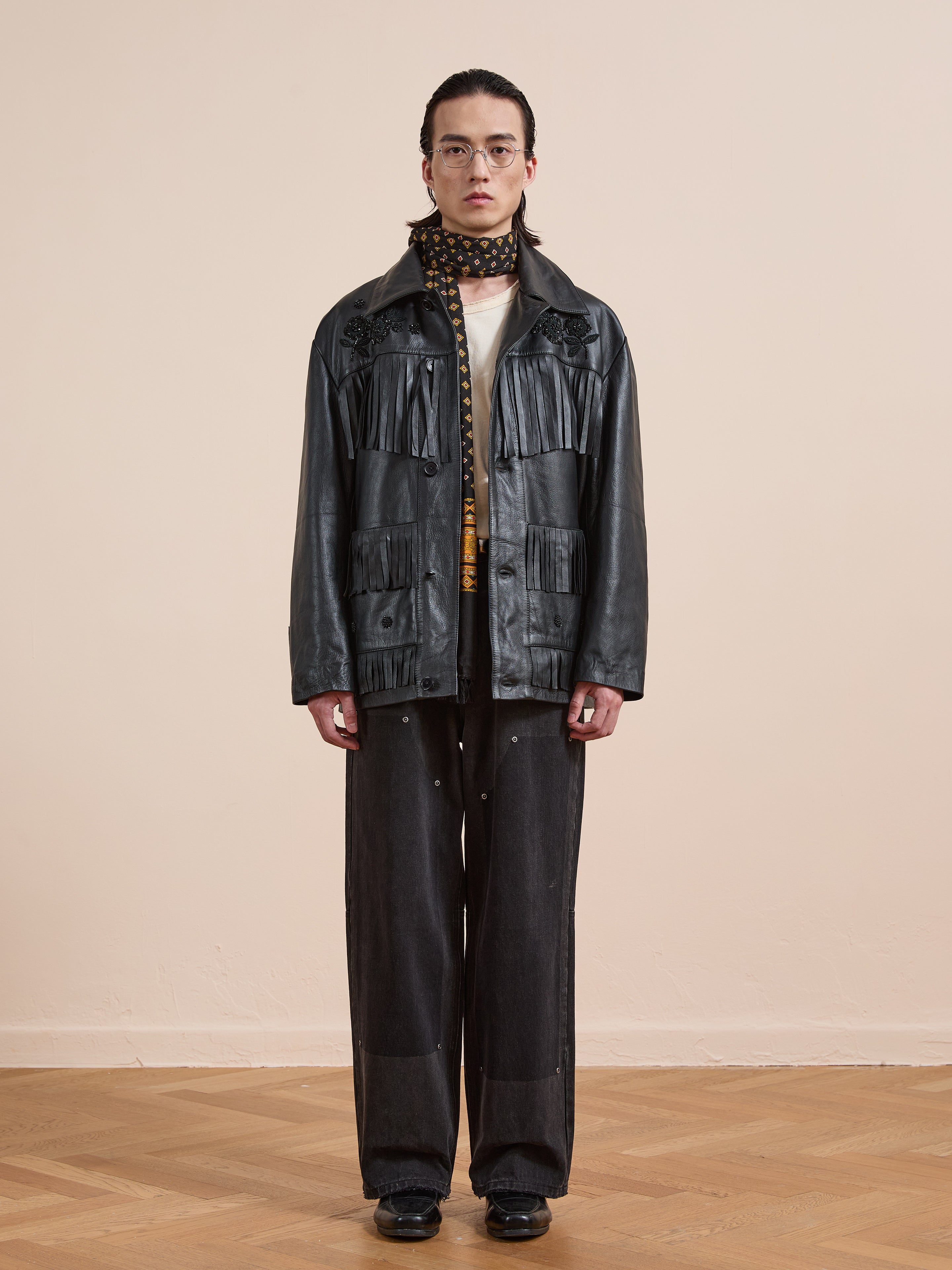 A person stands indoors wearing the FOUND Hand Embroidered Leather Fringe Jacket, paired with wide-leg black pants, a white shirt, glasses, and a patterned scarf.