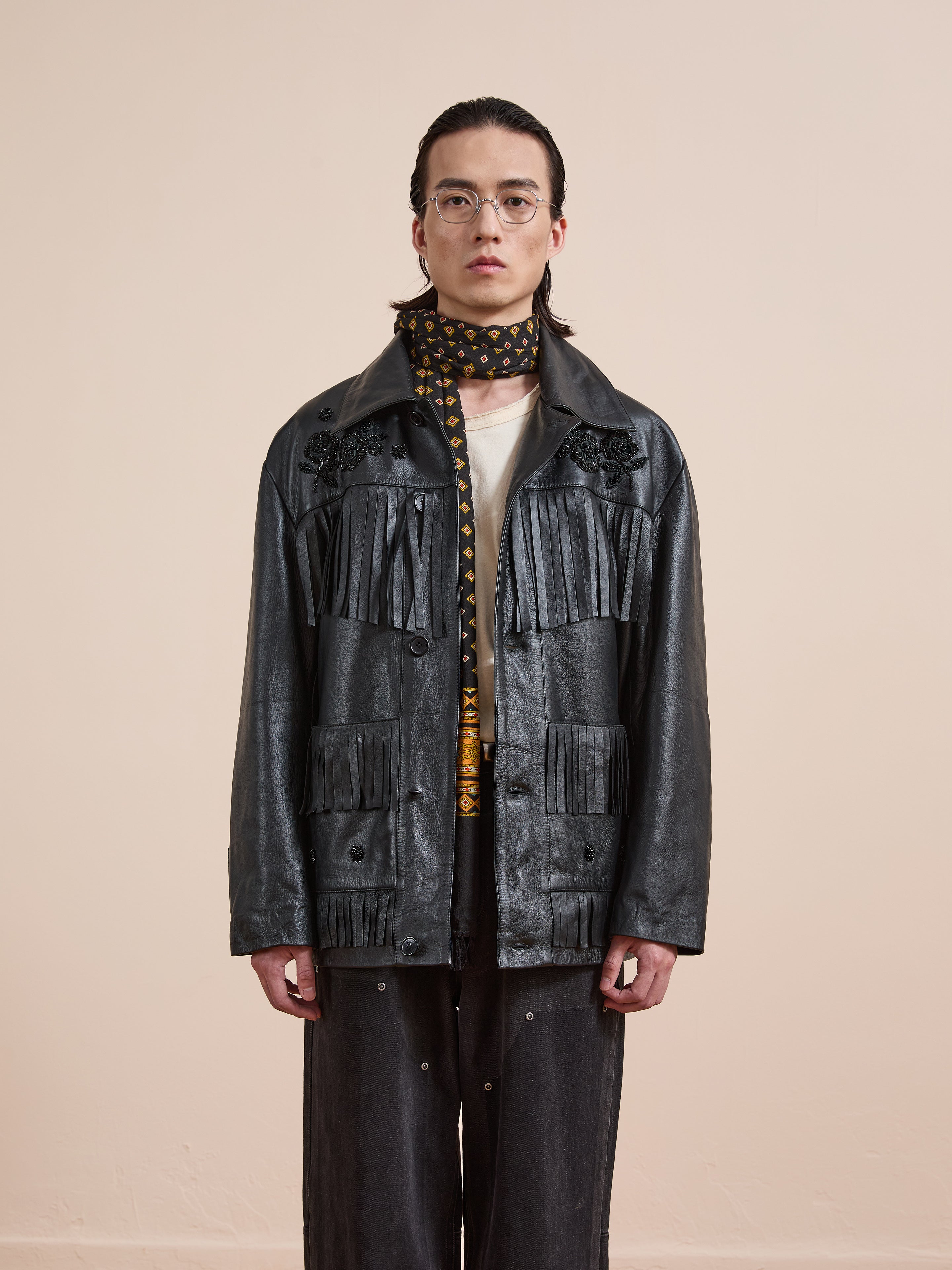 A person with long hair and glasses wears the FOUND Hand Embroidered Leather Fringe Jacket, a printed scarf, white shirt, and dark pants, standing against a plain light background.