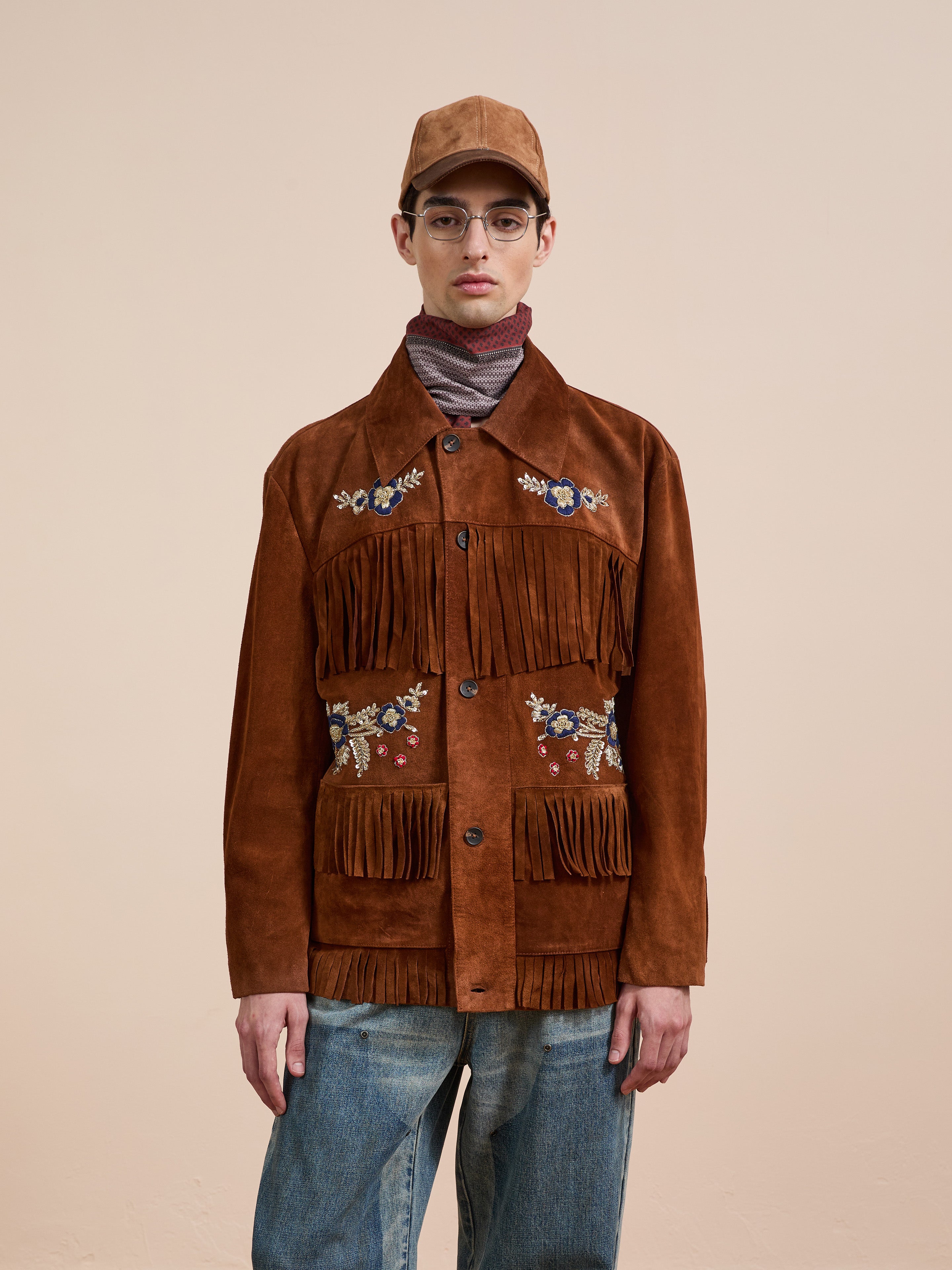 A person wearing the FOUND Hand Embroidered Suede Fringe Jacket, blue jeans, a brown cap, and glasses stands against a plain beige background.