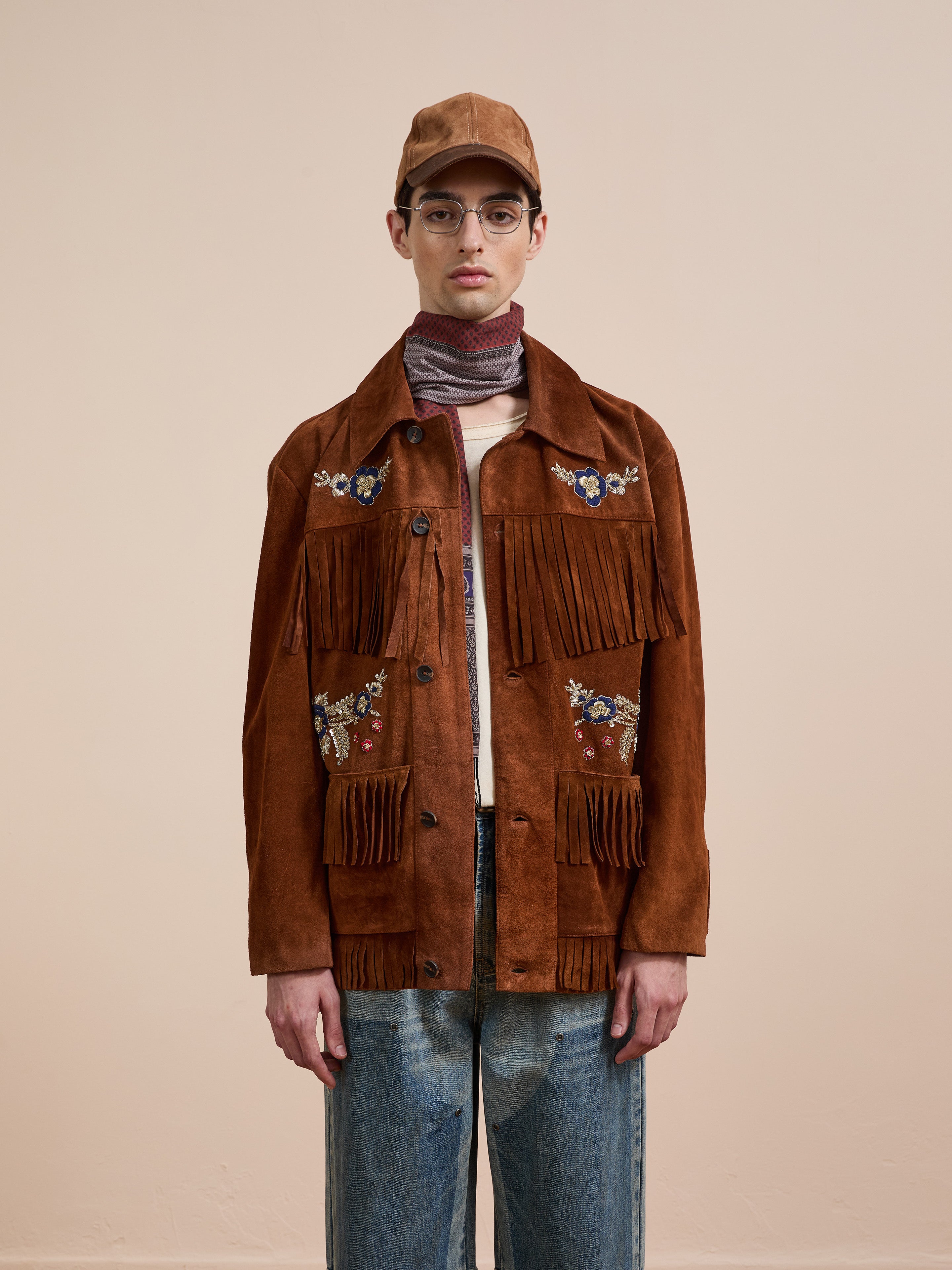 Wearing the FOUND Hand Embroidered Suede Fringe Jacket, blue jeans, a tan cap, glasses, and a striped scarf, a person stands against a plain background.