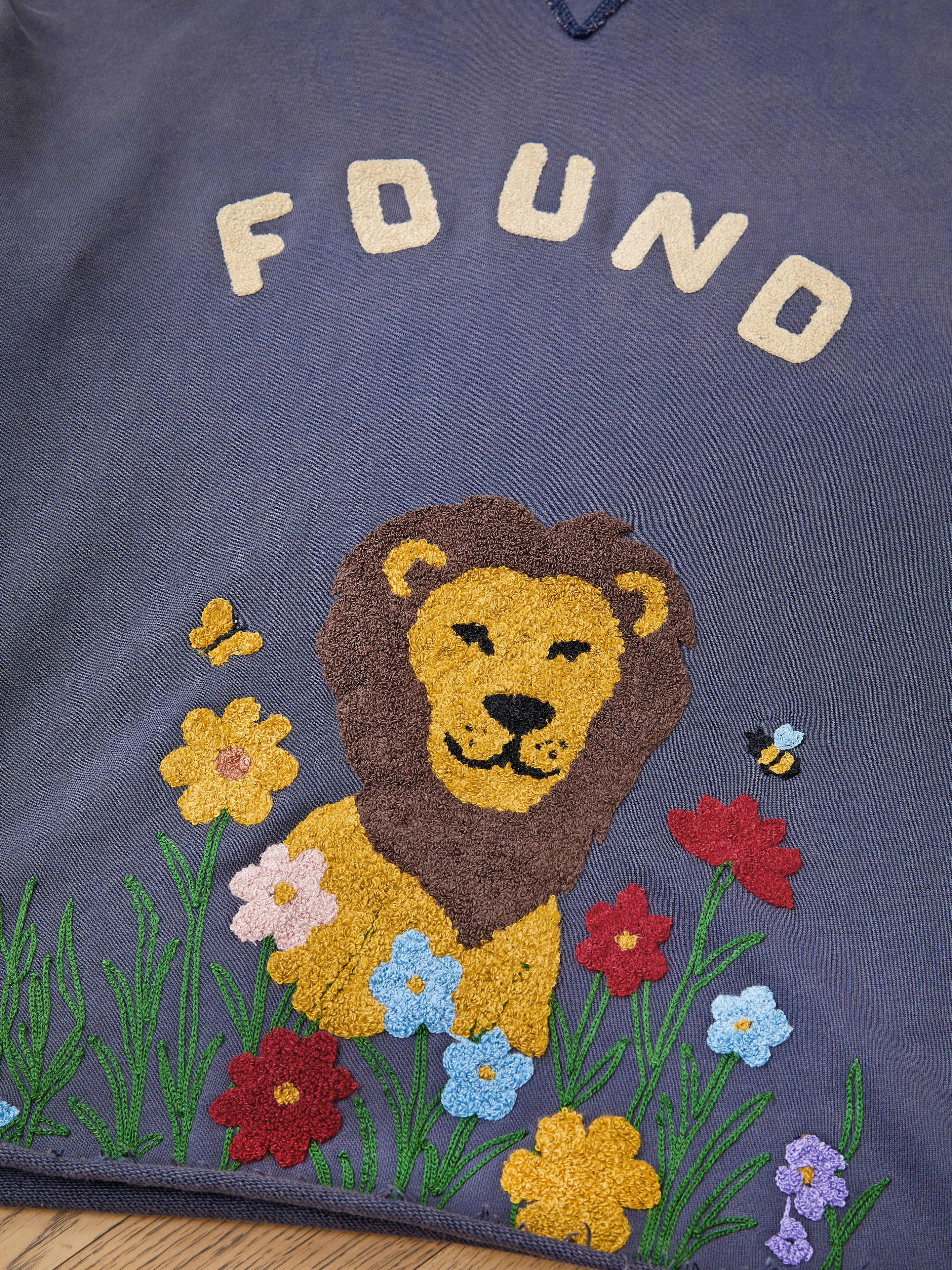 The Embroidered Wildlife Hoodie by FOUND features the brand name and a lion amid vibrant flowers, grass, and bees—offering unique wildlife-inspired charm in vintage French terry.