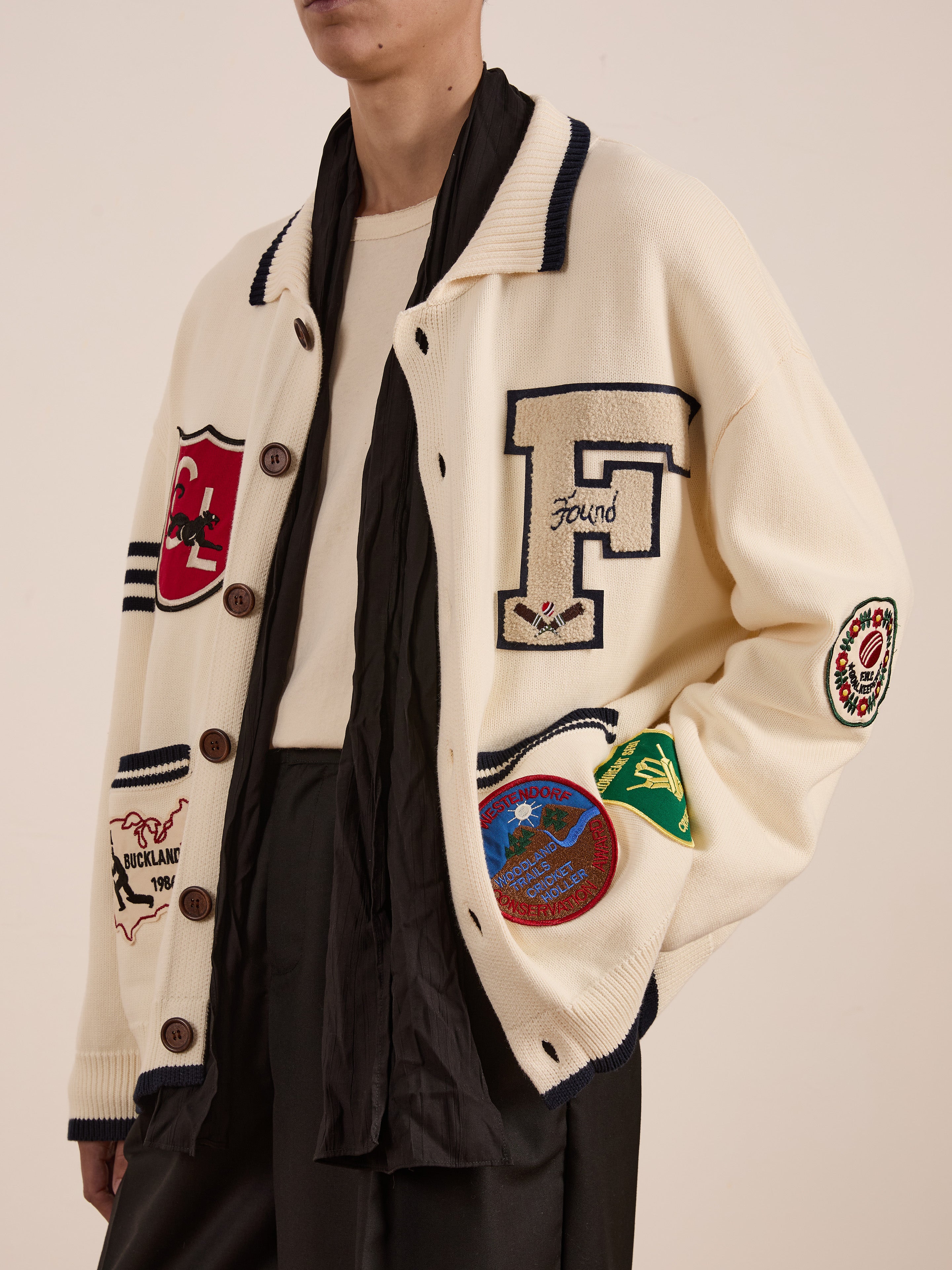 Exude collegiate nostalgia with Found's York Varsity Patch Collared Cardigan. This cream cardigan features various patches and a prominent "F" emblem, perfectly styled over a light shirt and dark pants.