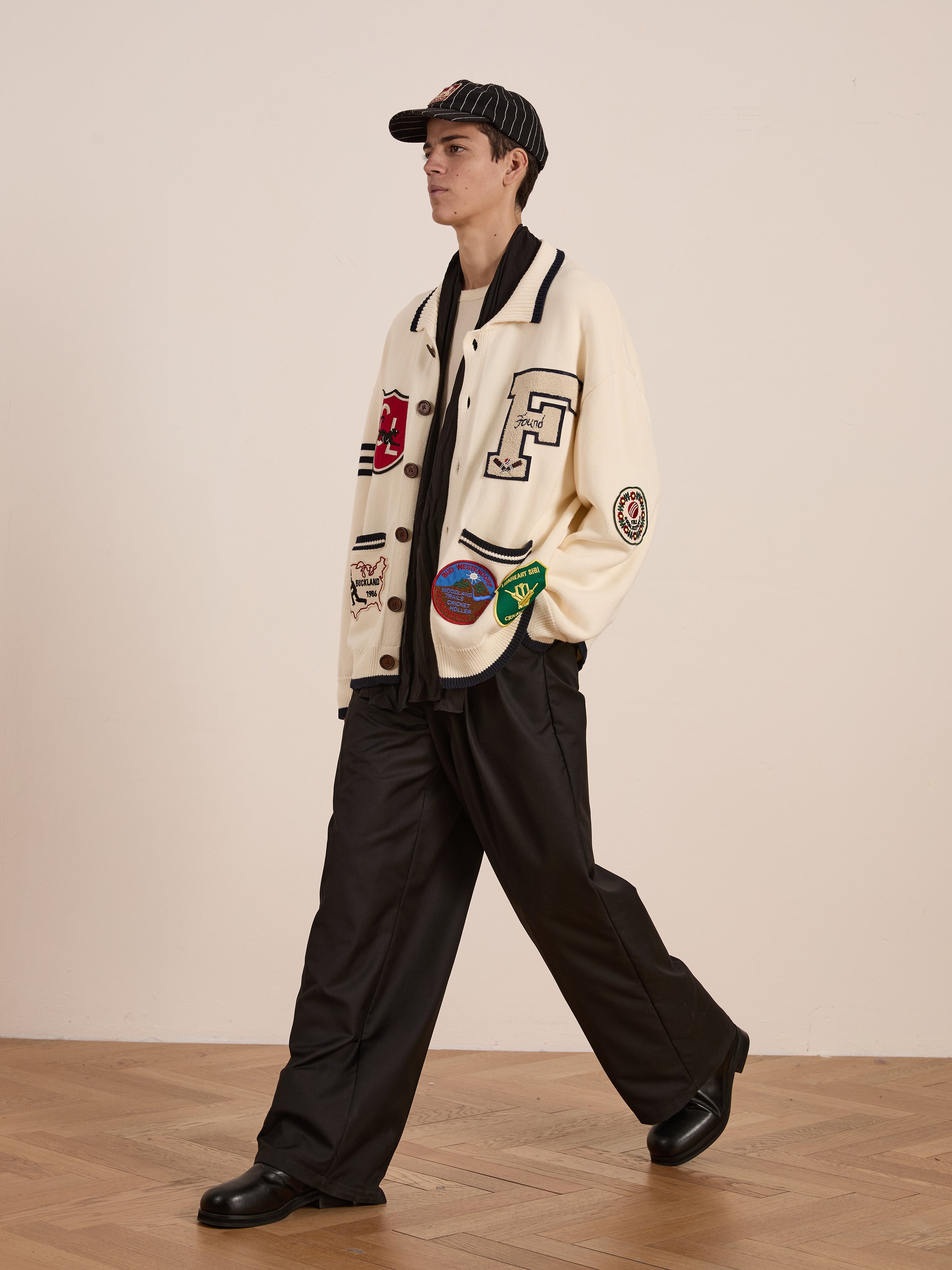 Wearing a cream York Varsity Patch Collared Cardigan by Found, black pants, and a striped cap, the person embodies a preppy aesthetic while walking on a wooden floor.