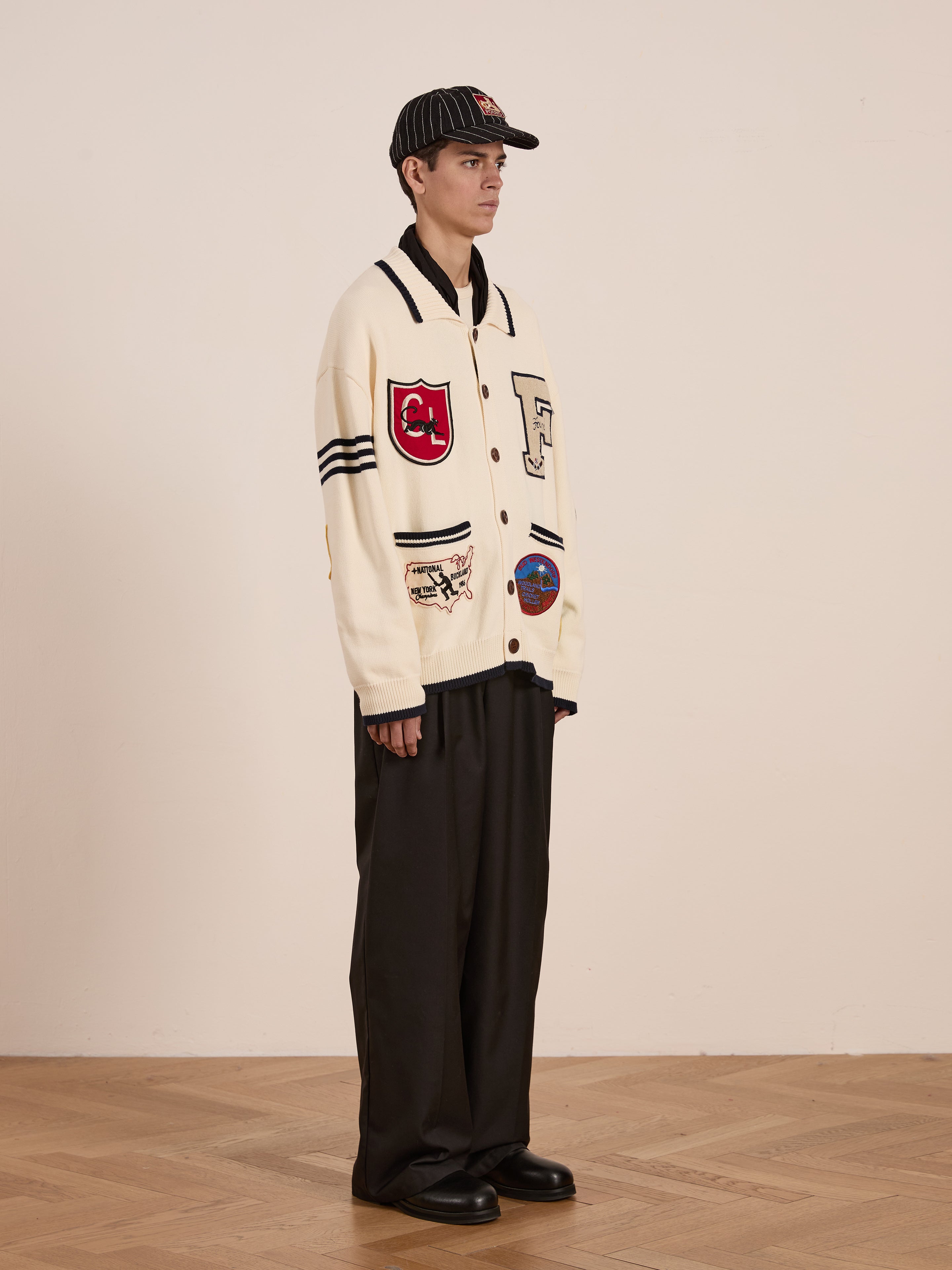 A person in a York Varsity Patch Collared Cardigan by Found exudes collegiate nostalgia, paired with black wide-leg pants, black shoes, and a striped cap, standing against a plain indoor background.