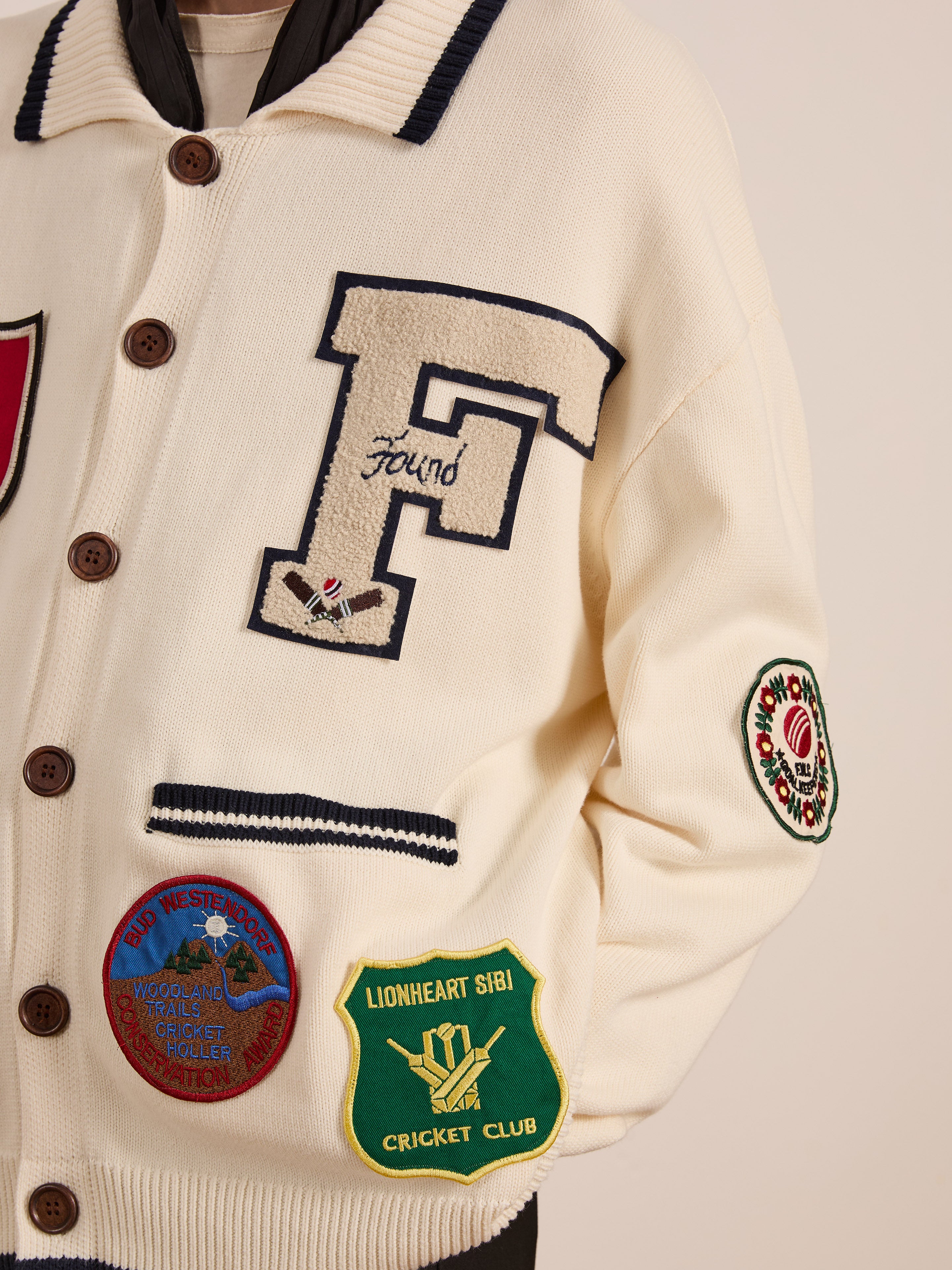 Wearing the York Varsity Patch Collared Cardigan by Found, featuring cream hues and embroidered patches like a prominent "F" and sports club logos, this piece captures a preppy vibe with collegiate nostalgia against a plain setting.