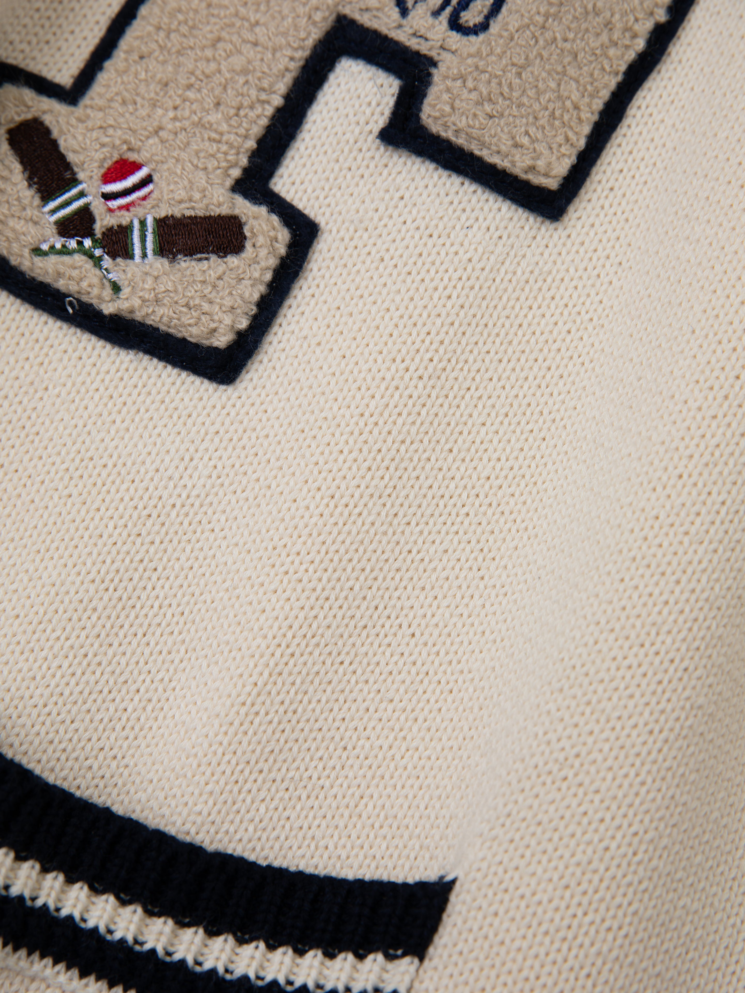 The York Varsity Patch Collared Cardigan by Found features a beige knit design with an embroidered 'H' and crossed cricket bats, reflecting collegiate nostalgia. Black and white striped detailing at the hem enhances its preppy style.