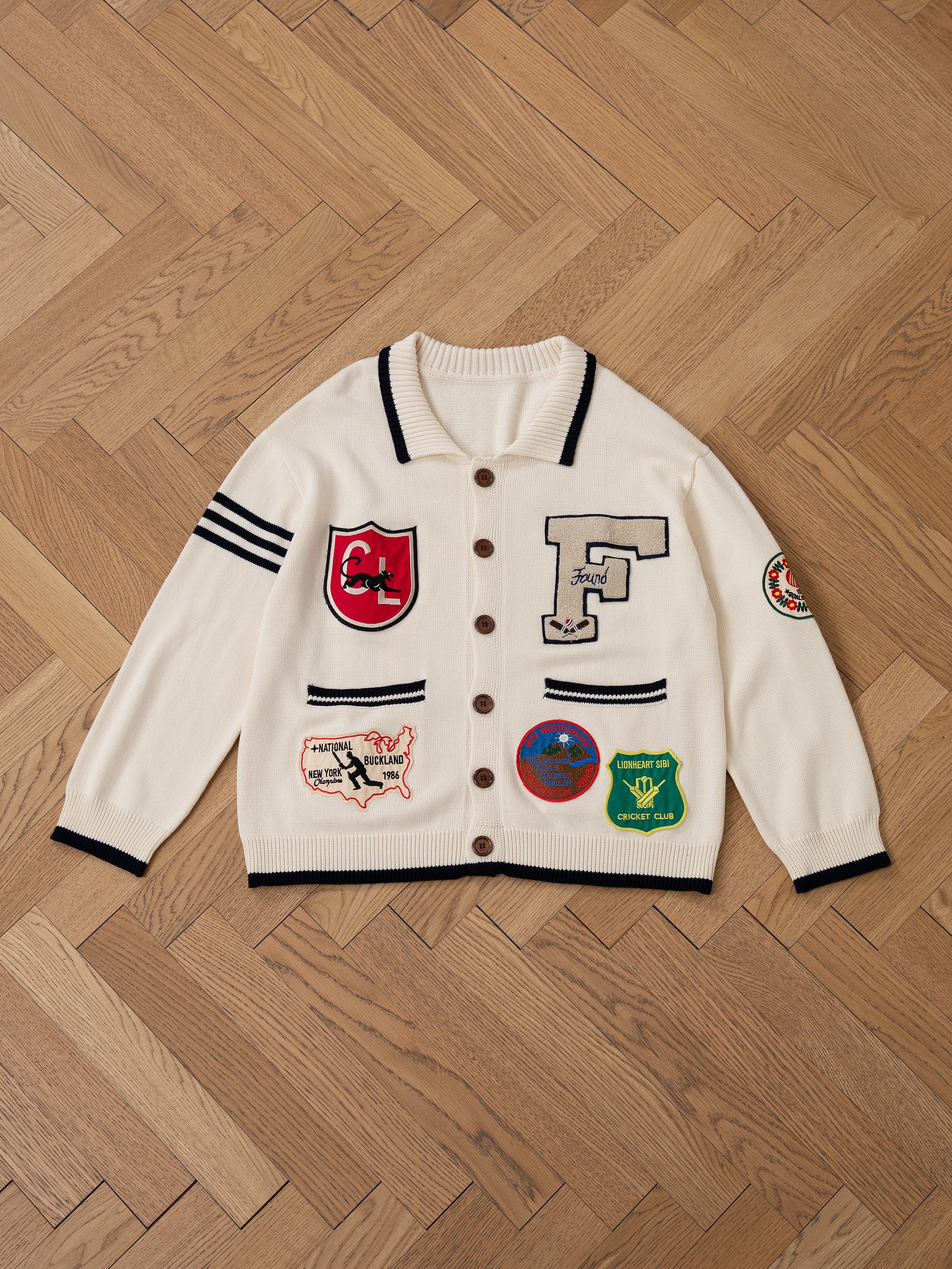 The York Varsity Patch Collared Cardigan by Found lies on a light wood floor, showcasing brown buttons and black trim. It has six assorted patches, including an "F," symbolizing a preppy style, while three black stripes on one sleeve add a touch of collegiate nostalgia.