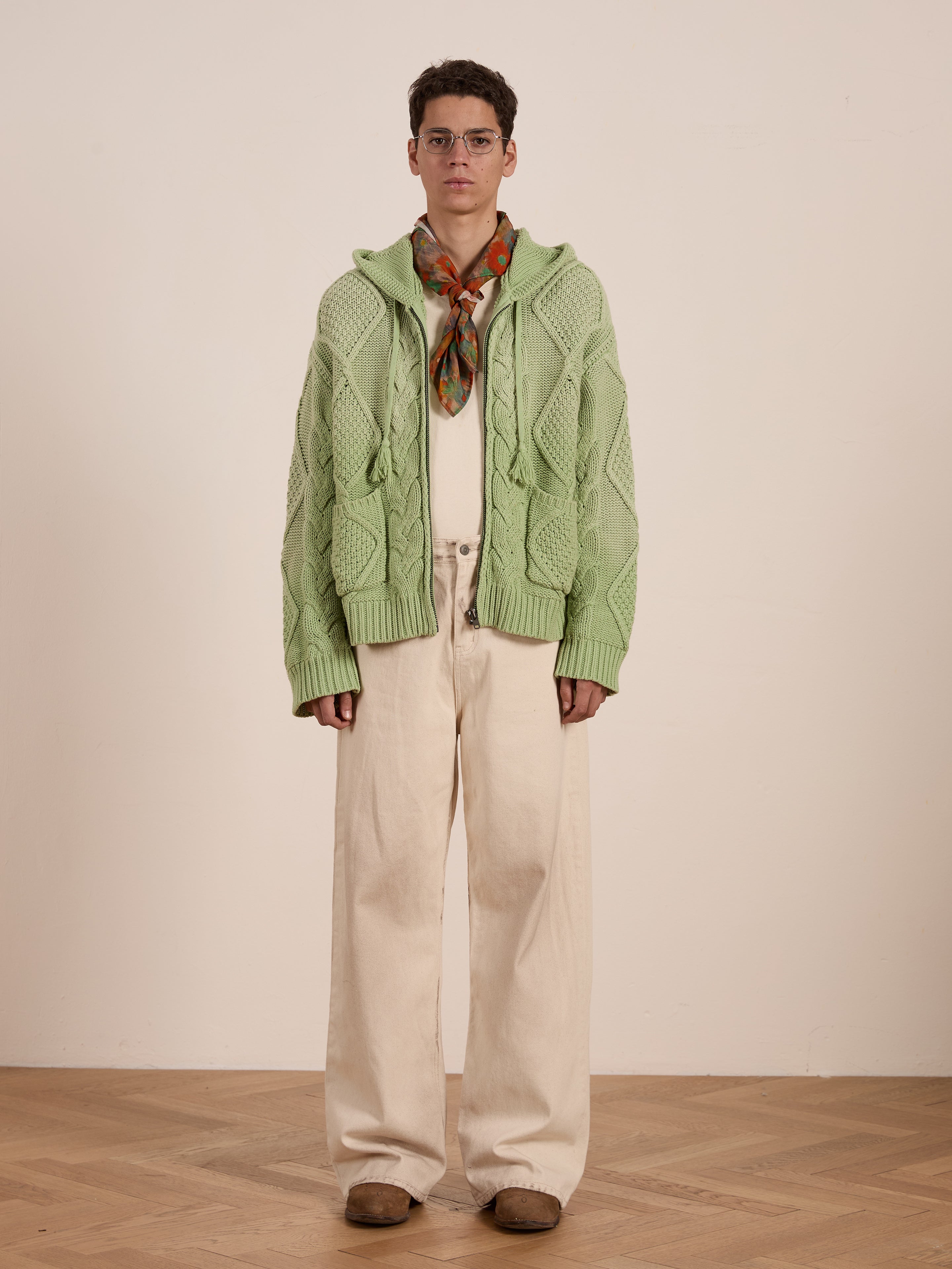 Wearing a Zip Up Cable Knit Hoodie by Found, a person with glasses stands indoors on a wooden floor. They're in beige wide-leg pants and brown shoes over a white shirt against a neutral background, exuding an effortlessly unisex relaxed fit.