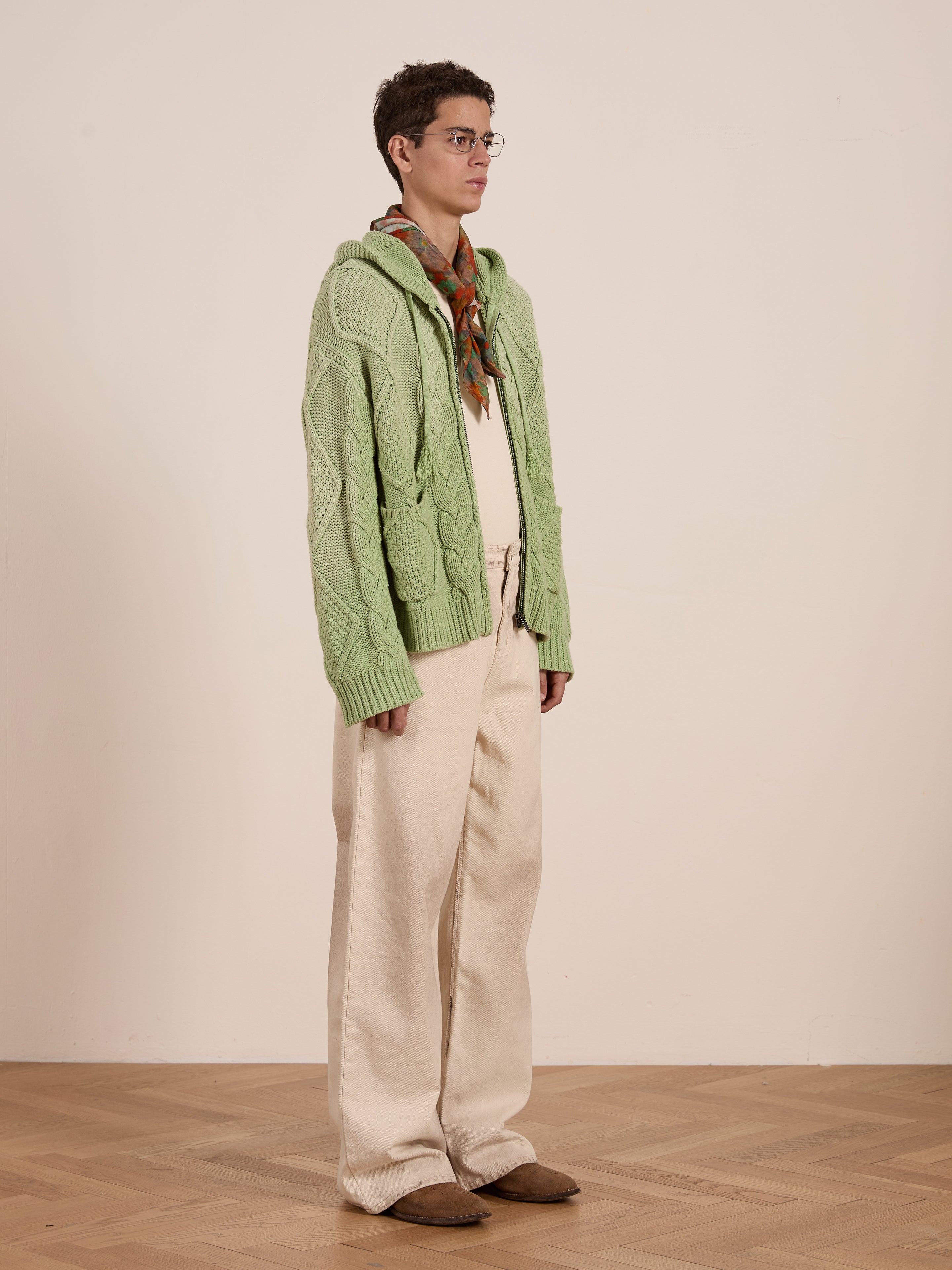 A person in a relaxed fit, oversized green Found Zip Up Cable Knit Hoodie and cream pants stands on a wooden floor, looking to the side.