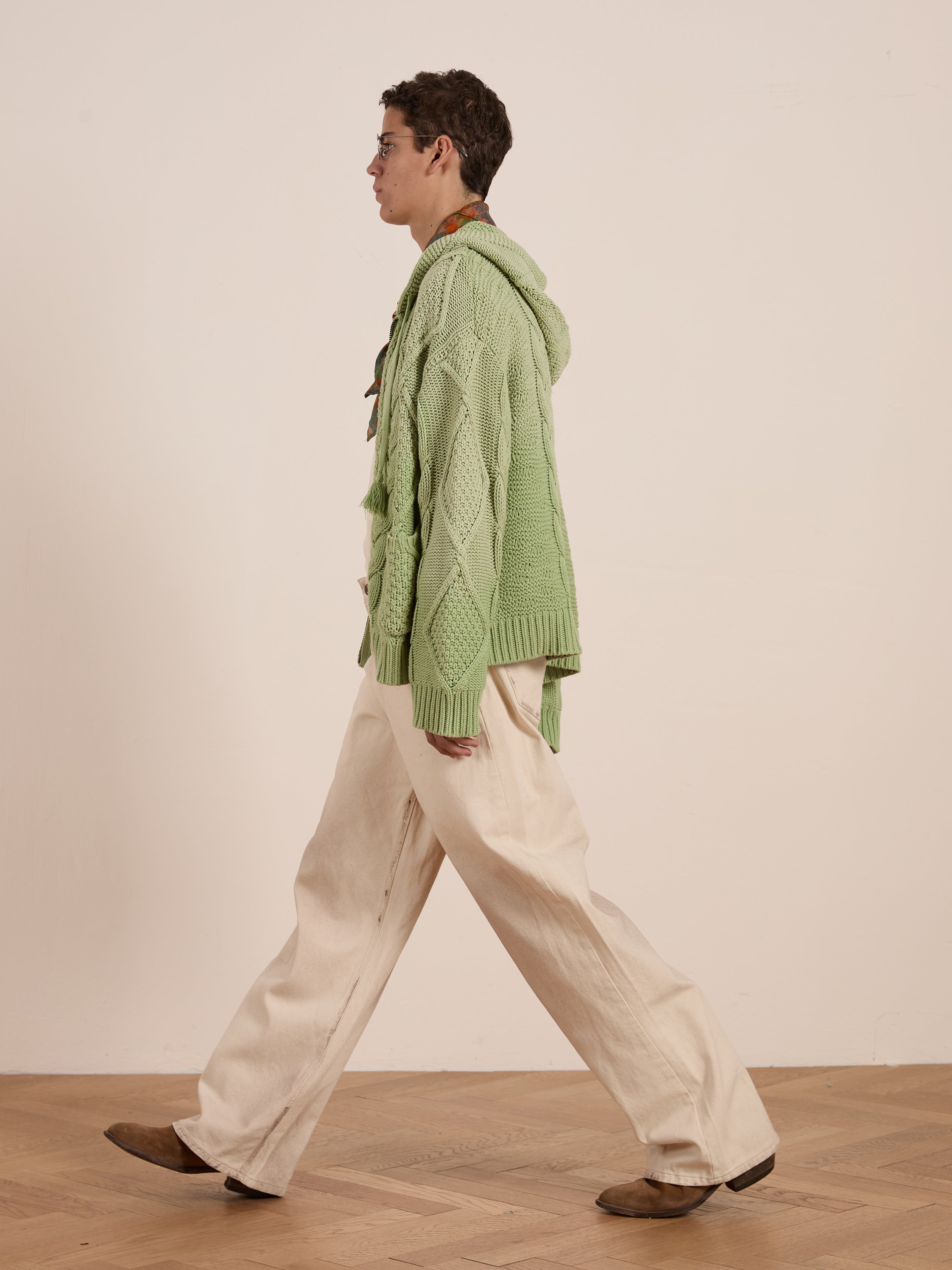 Wearing a Found Zip Up Cable Knit Hoodie crafted from a heavyweight cotton blend, the person in profile pairs the unisex relaxed-fit green top with beige wide-leg pants and brown shoes, confidently stepping across the wooden floor.