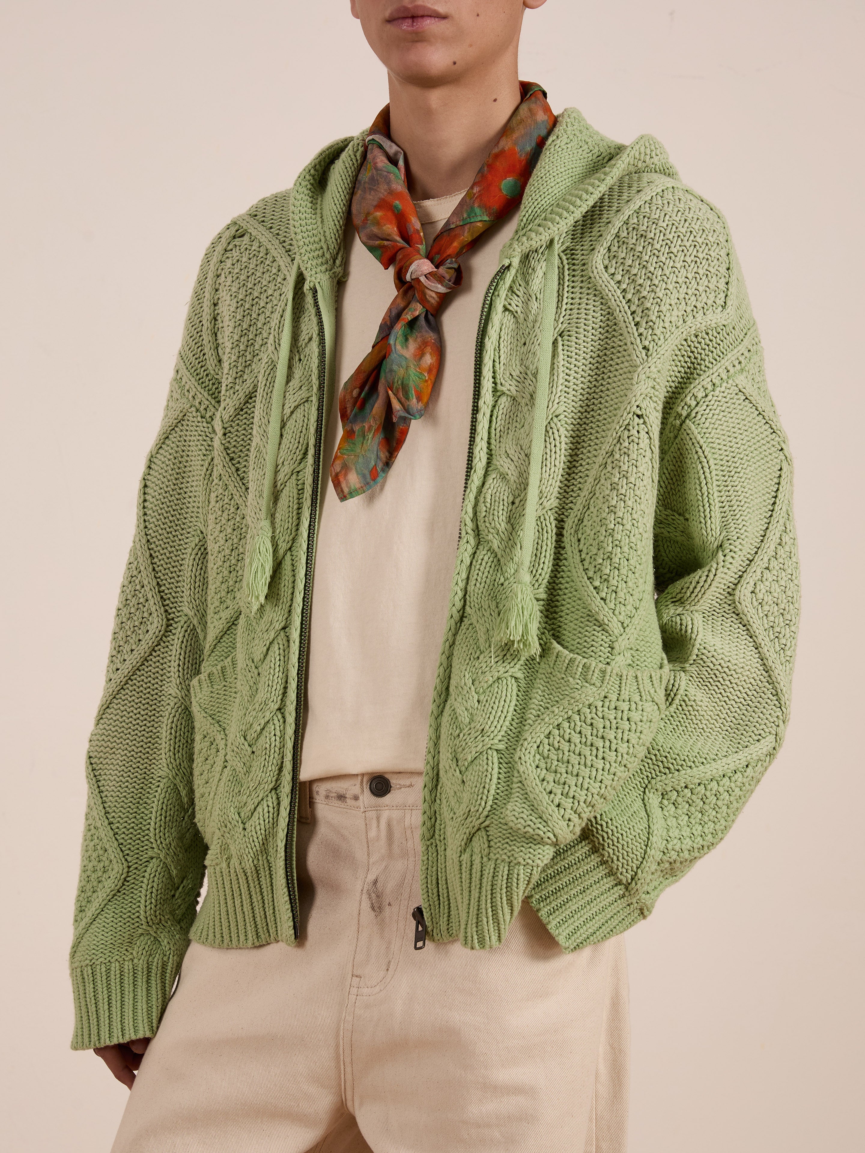 A person dressed in the cable knit hoodie from Found is wearing a green zip-up version with beige pants and a colorful scarf. The unisex relaxed fit ensures comfort and style, crafted from a heavyweight cotton blend against a neutral background.