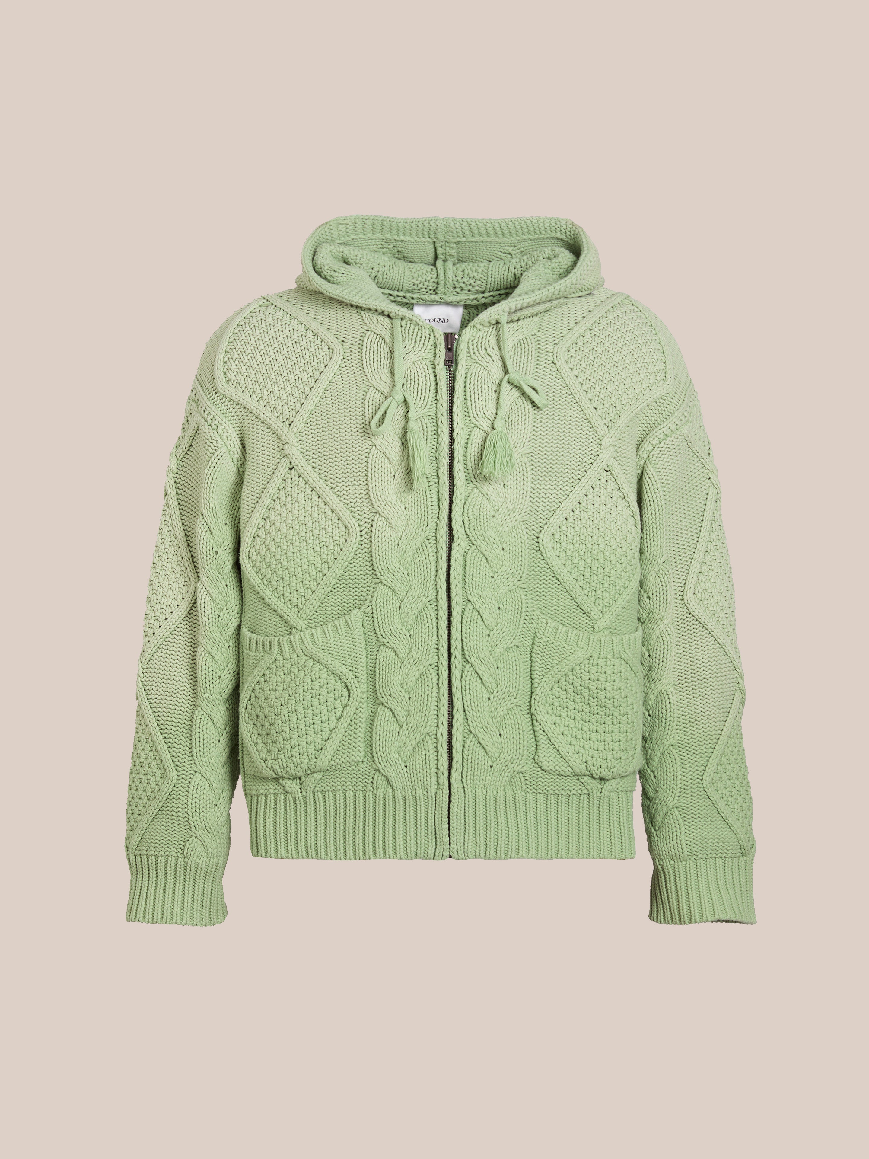 The Zip Up Cable Knit Hoodie by FOUND is a green unisex cotton blend with a textured pattern, zip-up front, and drawstring hood, set against a beige background in a relaxed fit.