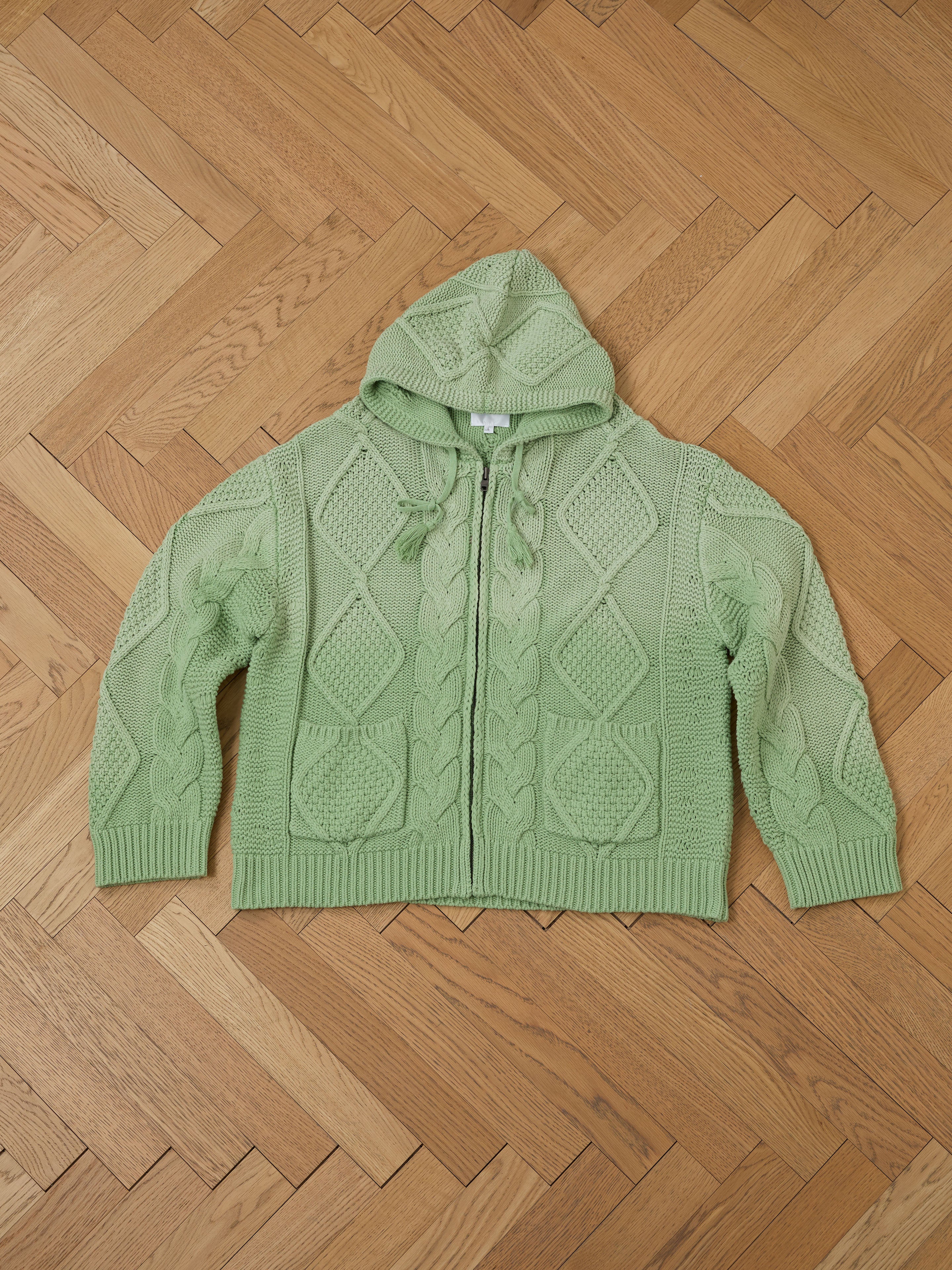 The Found Zip Up Cable Knit Hoodie, a green heavyweight cotton blend with a relaxed fit, is displayed flat on a wooden herringbone floor.