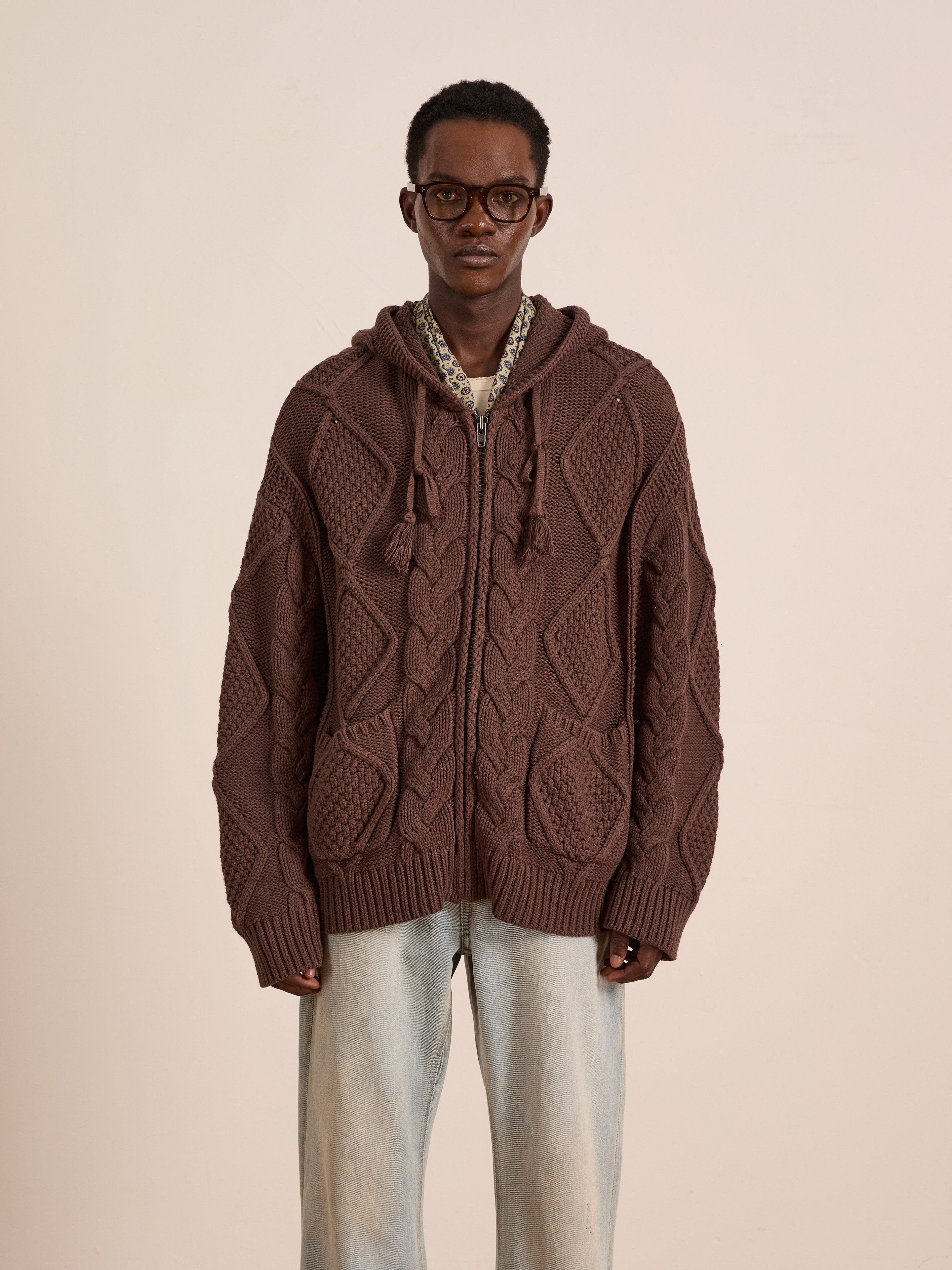 A person wearing glasses, a FOUND Zip Up Cable Knit Hoodie, a patterned shirt, and light blue jeans stands against a plain beige background.