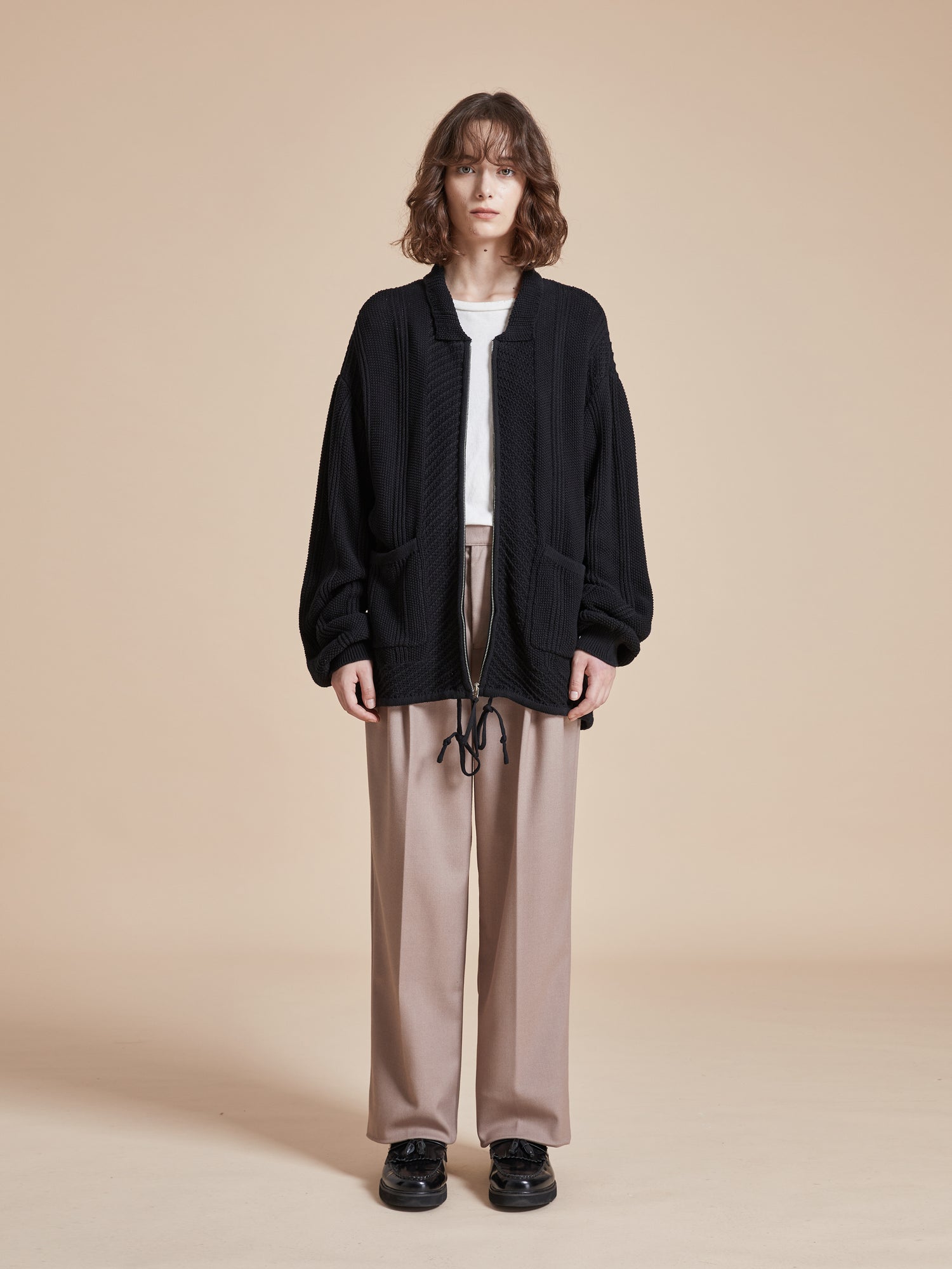 A model wearing a black jacket and beige pants with Found's Zip Up Panel Knit Ribbed Sweater.