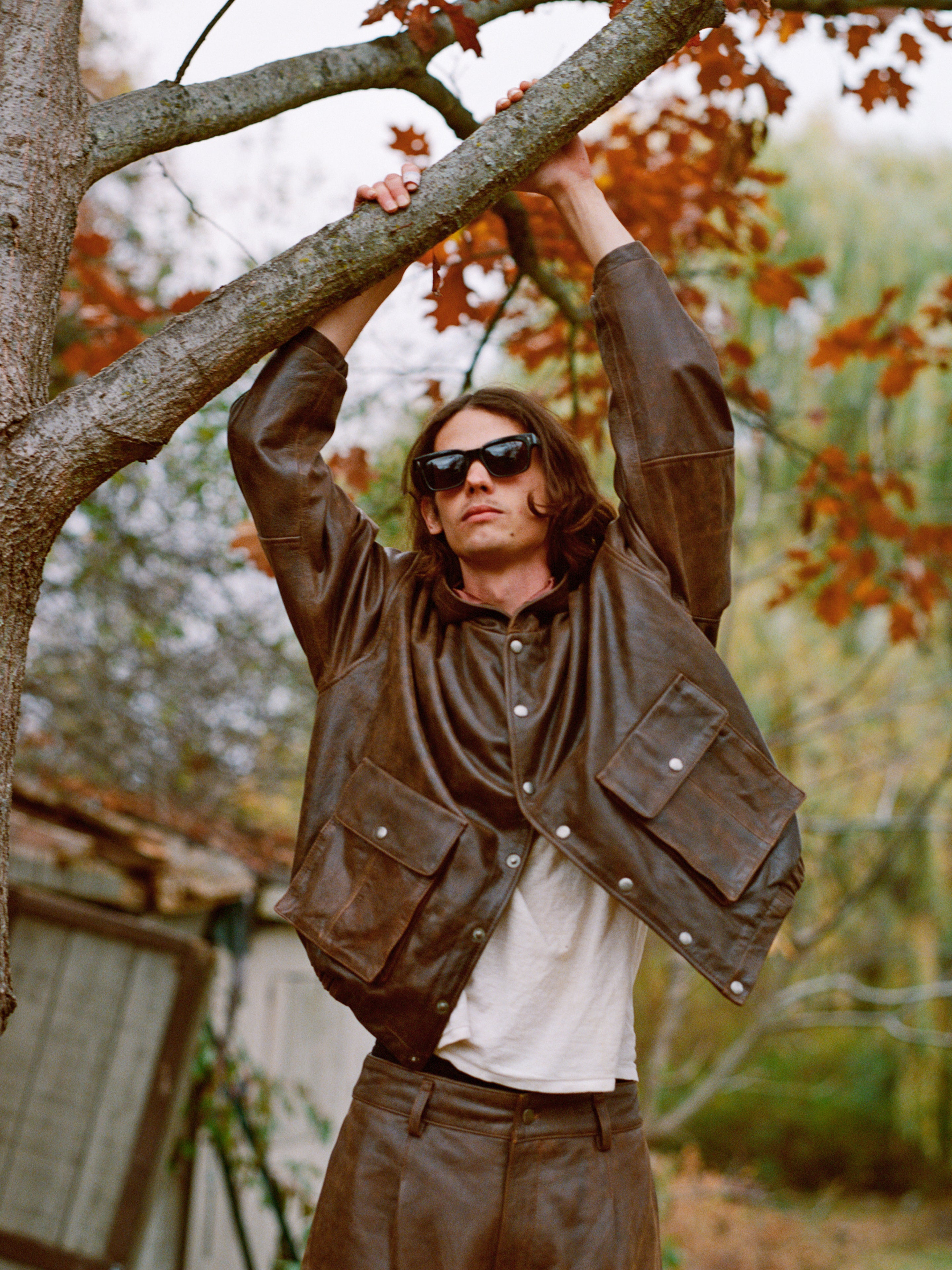 Wearing Dusk Distressed Genuine Leather Hoodie by FOUND, a person in sunglasses hangs from a tree branch with vibrant autumn leaves as the backdrop.