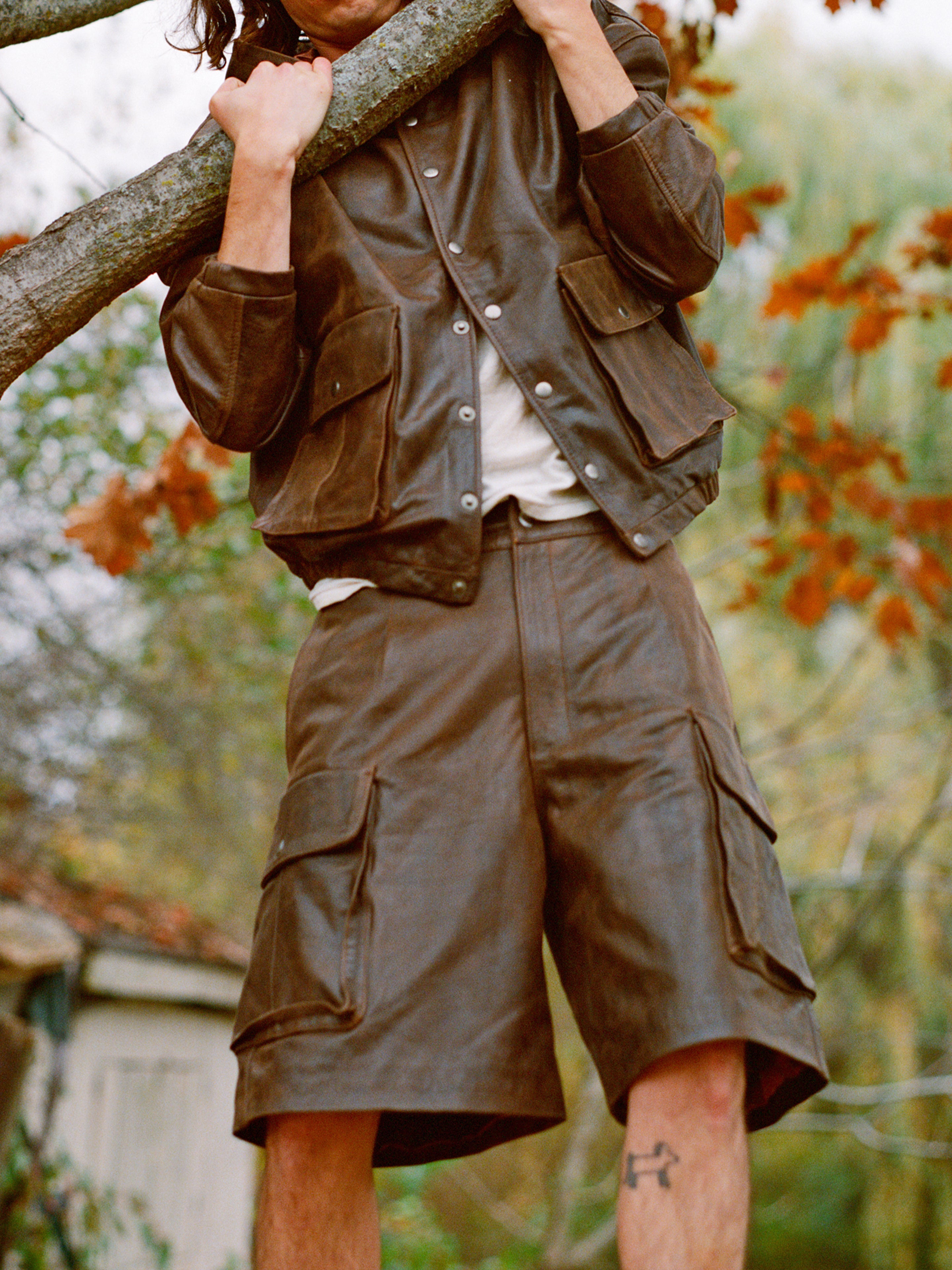 A person in a FOUND Dusk Distressed Genuine Leather Hoodie and shorts clings to a tree branch, surrounded by the vibrant hues of autumn leaves.