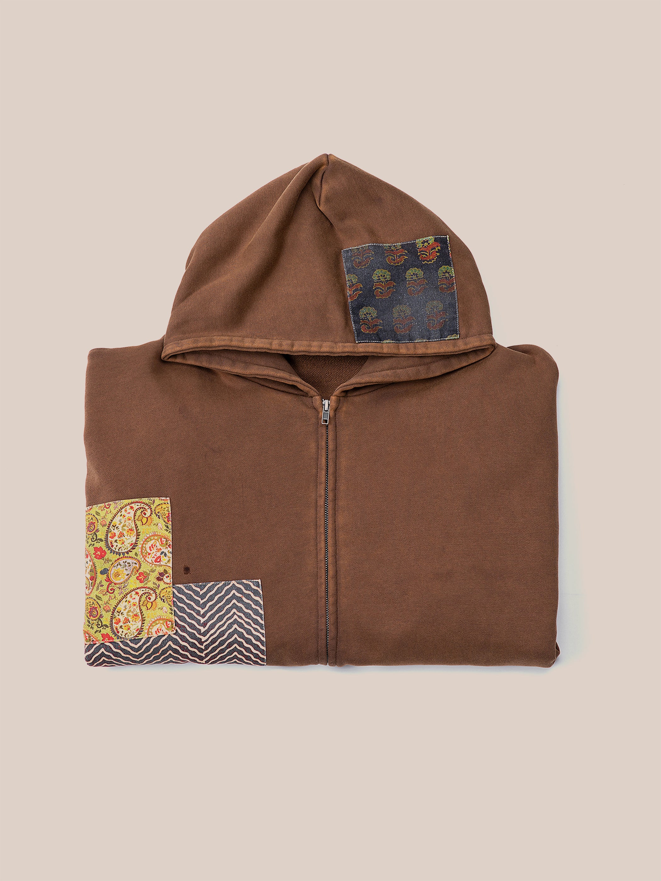 The Tapestry Patchwork Zip Up Hoodie by FOUND is an oversized brown hoodie with a front zipper, showcasing multicolored patch designs on the hood and lower left side, inspired by South Asian tapestry.