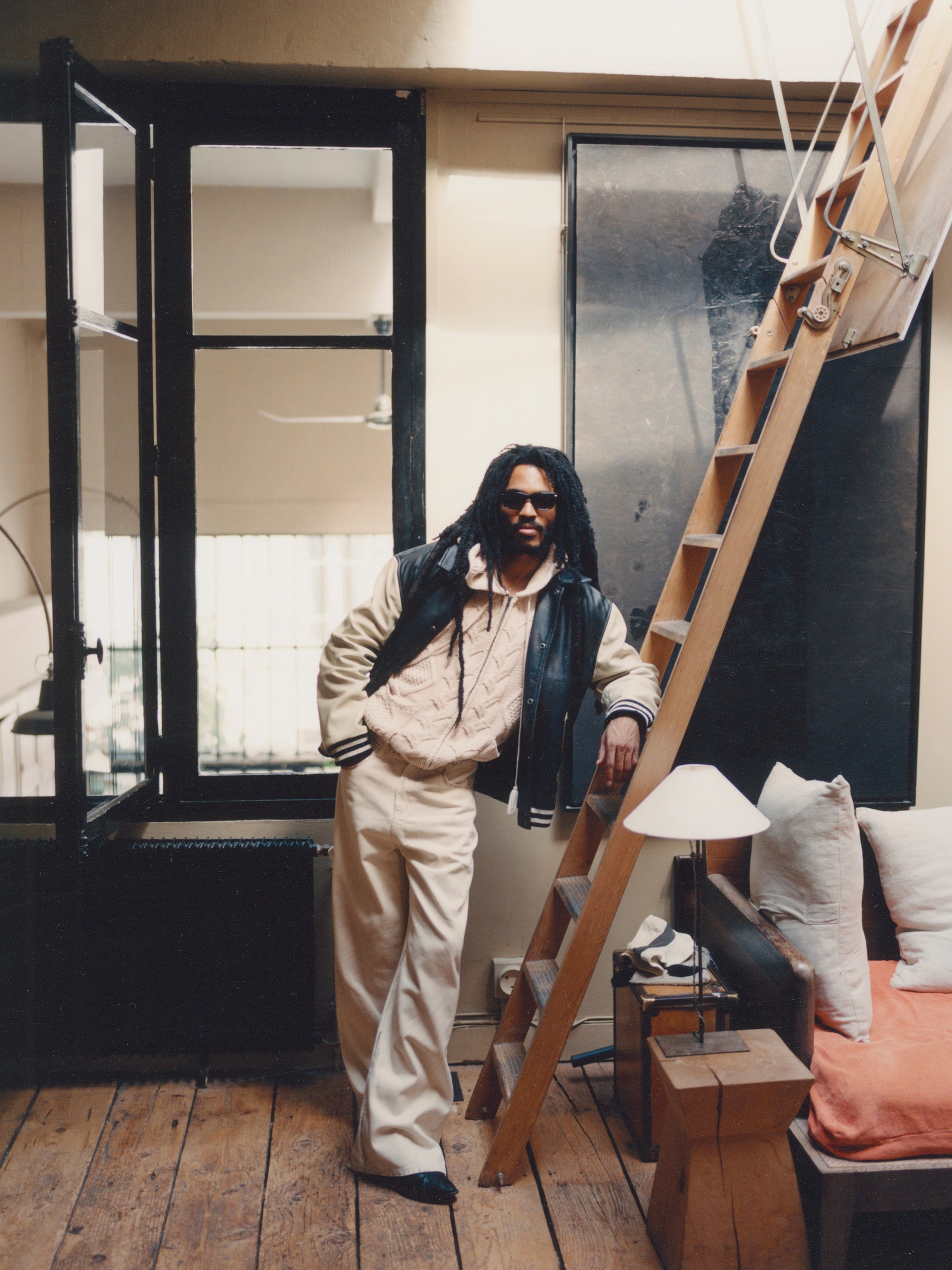 A person wearing sunglasses, beige pants, and a FOUND Logan Distressed Leather Varsity Jacket with satin lining stands indoors, leaning on a wooden ladder. An open window behind them reveals a room with a lamp, couch, and cushions in the background.