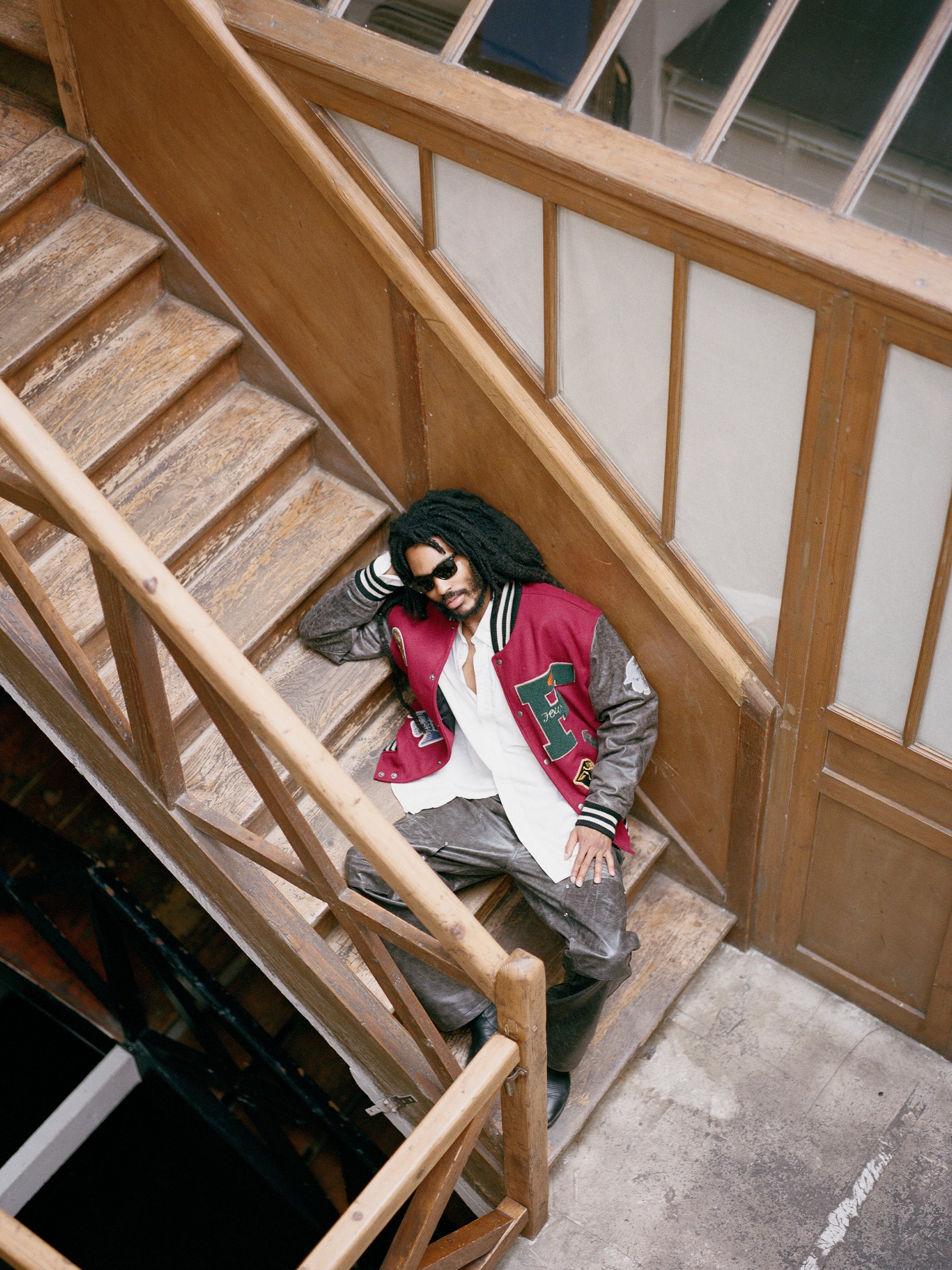 A person wearing a Found Crimson Letterman Jacket and sunglasses is reclining on wooden stairs in an indoor setting, exuding a vintage vibe.