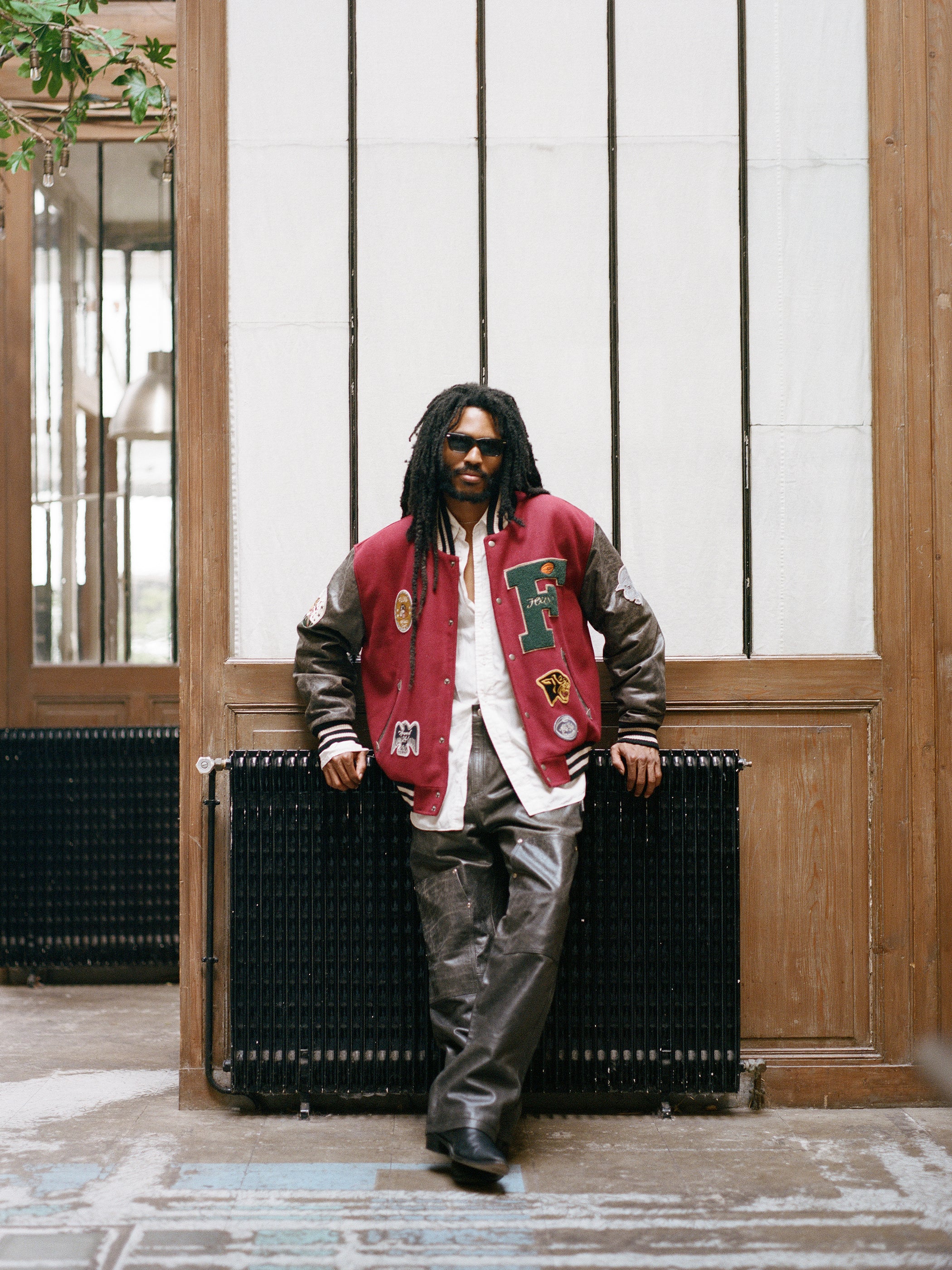 A person with dreadlocks, donning sunglasses, a Crimson Letterman Jacket by Found made from ethically sourced materials, and leather pants leans against a black radiator in a wooden interior space.