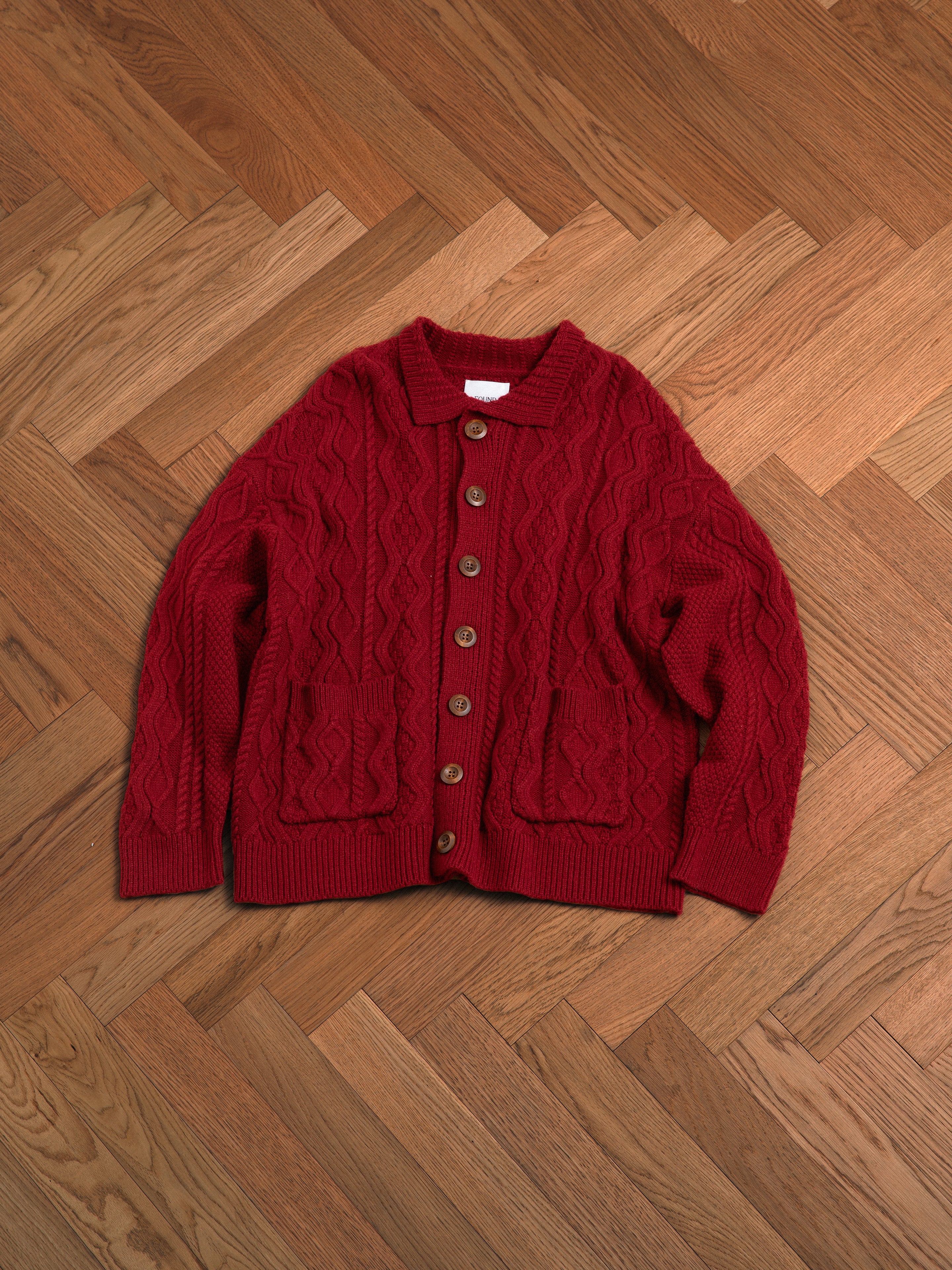 A red-hued yarn, Found Parsidan Cable Knit Cardigan sitting on a wooden floor.