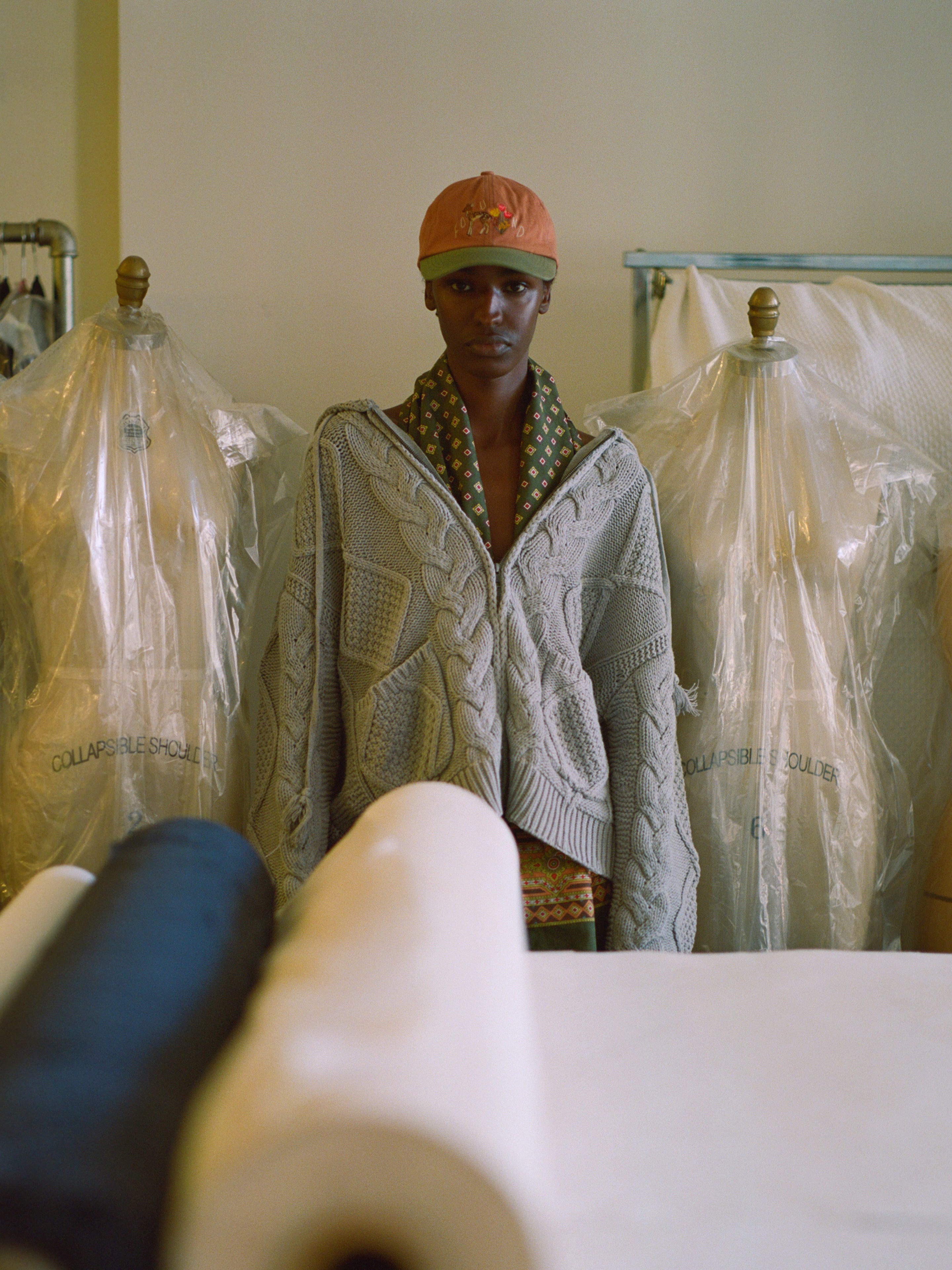 A person in a Found Zip Up Cable Knit Hoodie and orange cap stands in a room with garment bags on dress forms and rolls of fabric in the foreground.