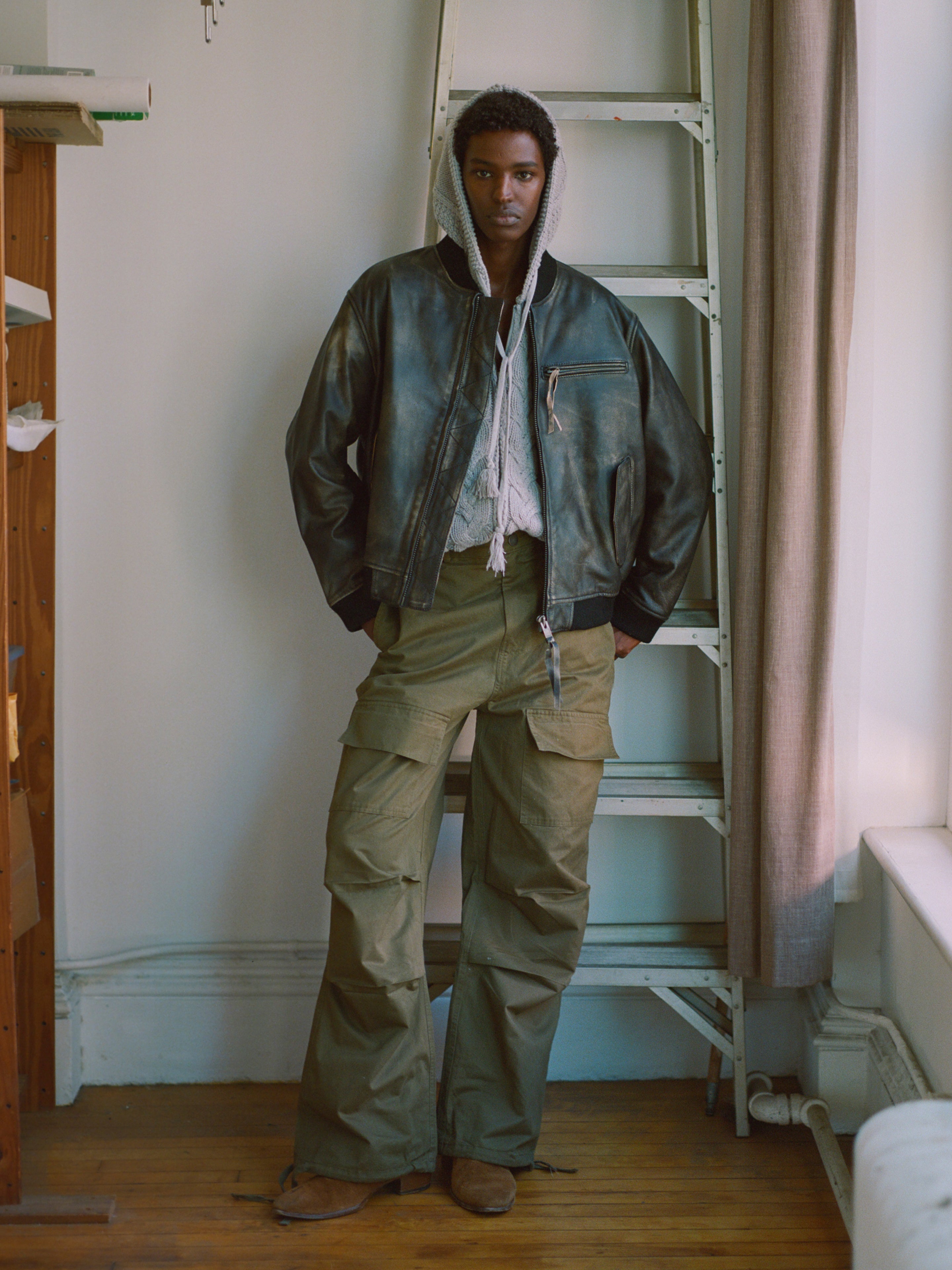 A person stands indoors wearing a black leather jacket, a Found Zip Up Cable Knit Hoodie, and green cargo pants with their hands in their pockets. A ladder and a curtain are visible in the background.