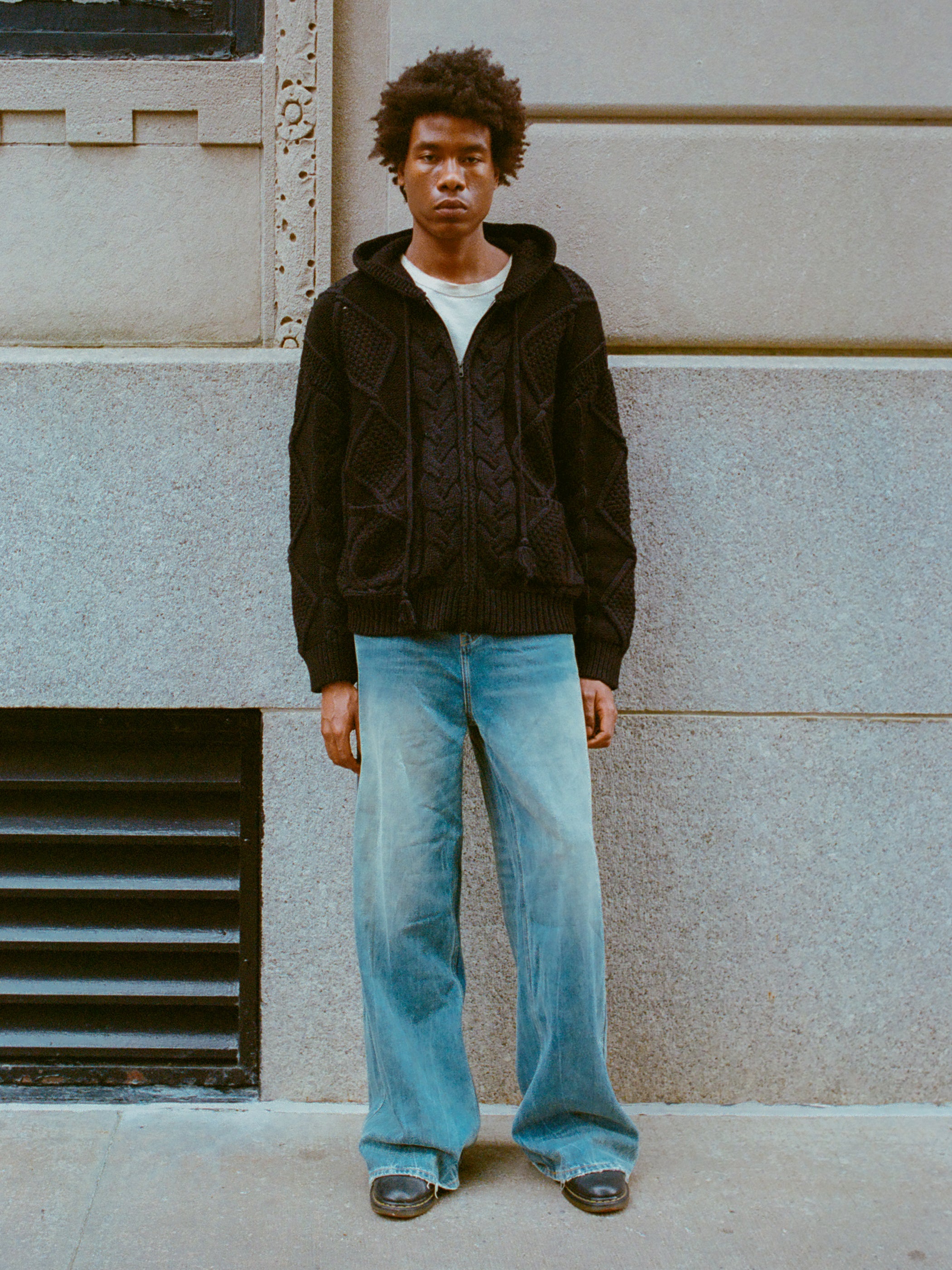 A person with an afro hairstyle stands against a light gray building wall, wearing the Found Zip Up Cable Knit Hoodie and faded blue jeans.