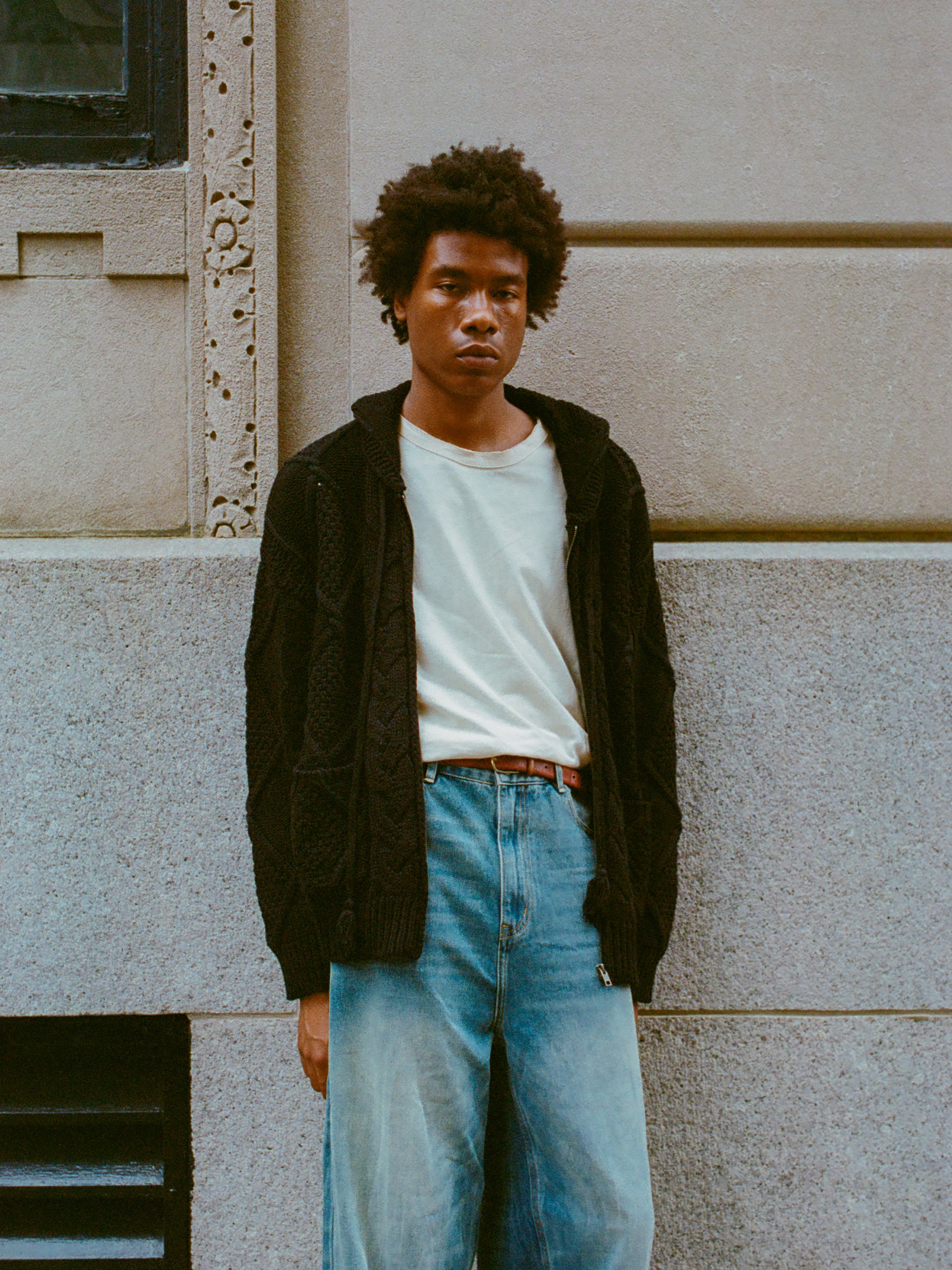 A person with short curly hair stands against a stone wall, wearing a white T-shirt, a heavyweight cotton blend black Zip Up Cable Knit Hoodie from Found, and light blue jeans, creating a cozy look and feel.