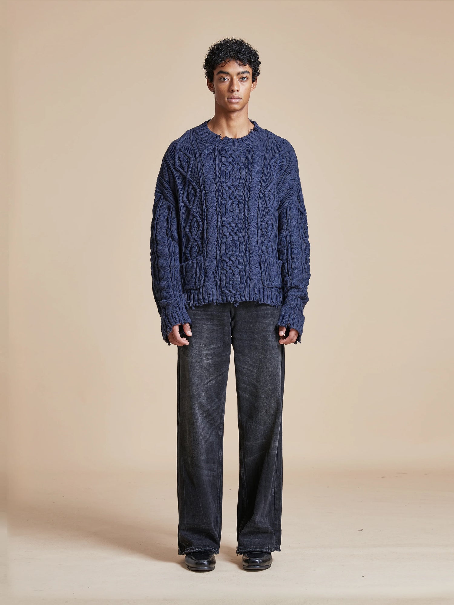 A man donning an Astral Distressed Cable Knit Sweater from Found in softness and warmth, paired with black pants.