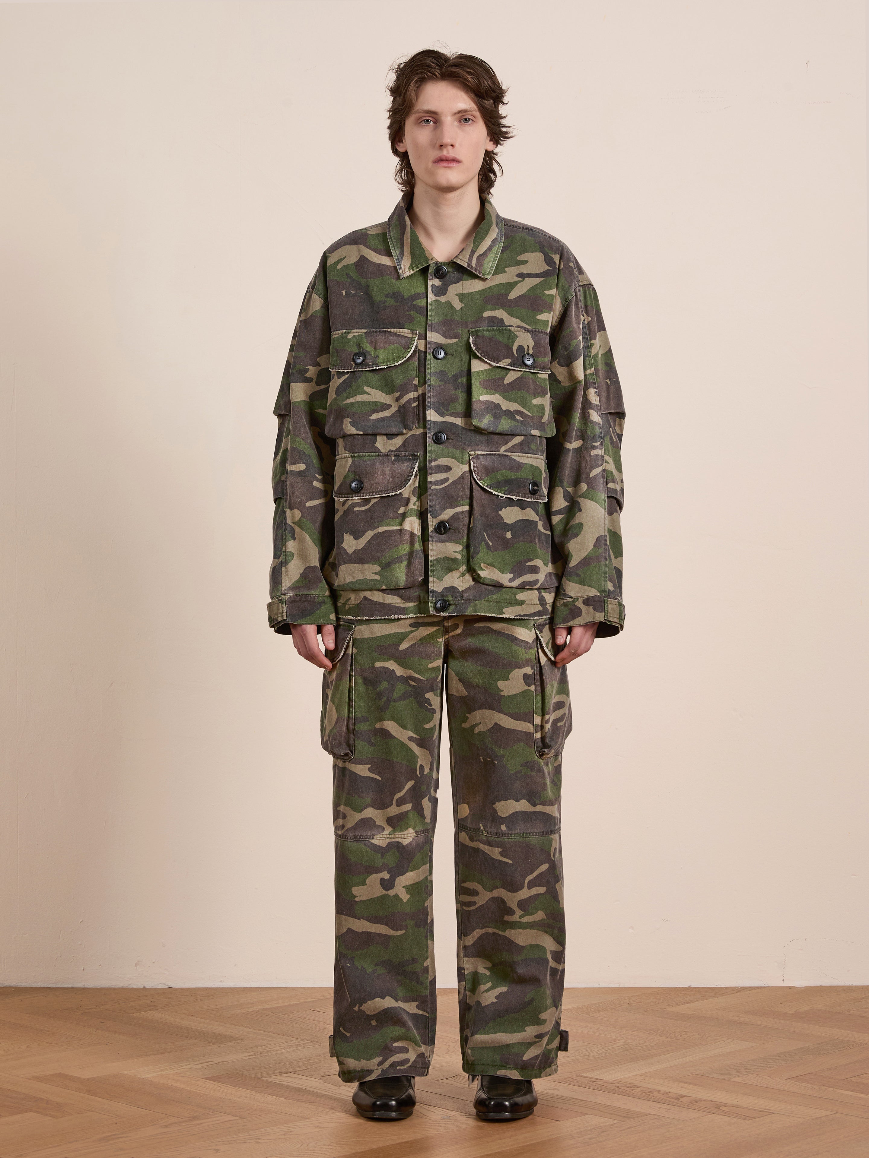 Indoors on a wooden floor, a person dons the FOUND Camouflage Twill Utility Cargo Jacket and pants, showcasing a vintage style.