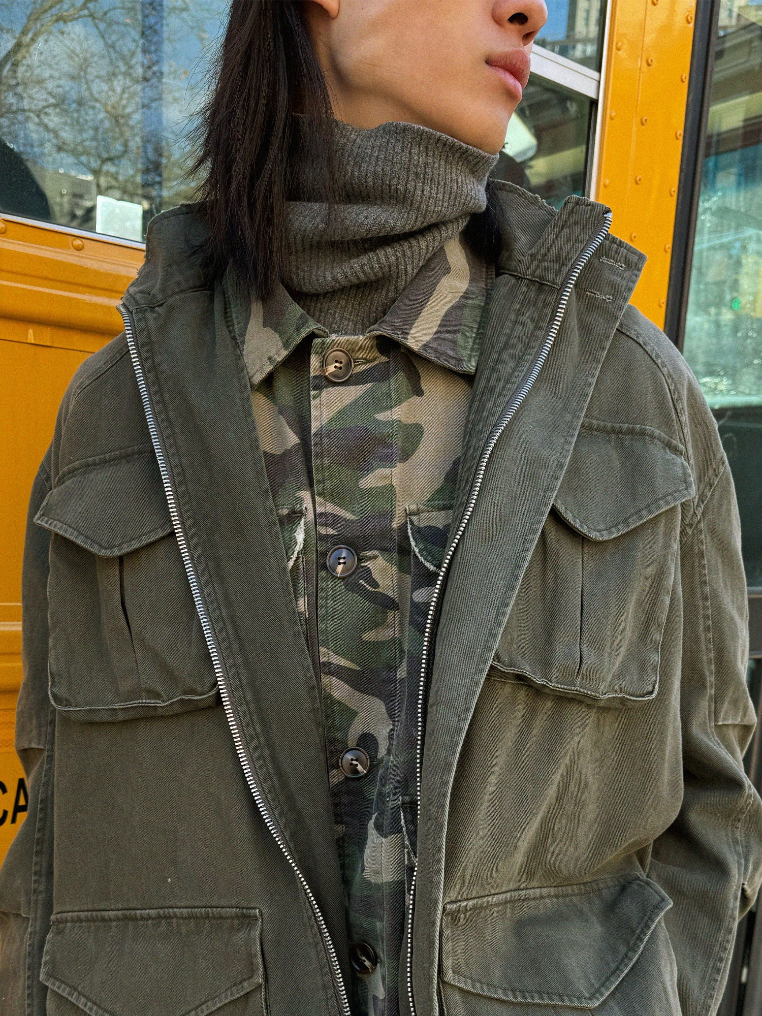 A person confidently stands in front of a yellow vehicle wearing FOUND's Camouflage Twill Utility Cargo Jacket over a camo shirt and a vintage-look gray turtleneck.