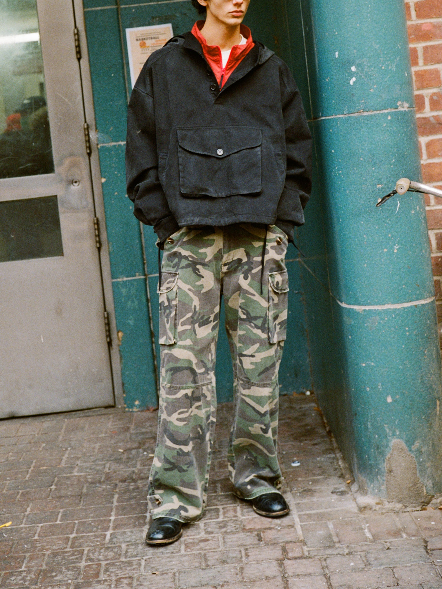 A person in a FOUND vintage black Washed Twill Pocket Anorak Jacket with a red collar and camouflage cargo pants stands outside near a green pillar and gray door, embodying unisex relaxed fit style.