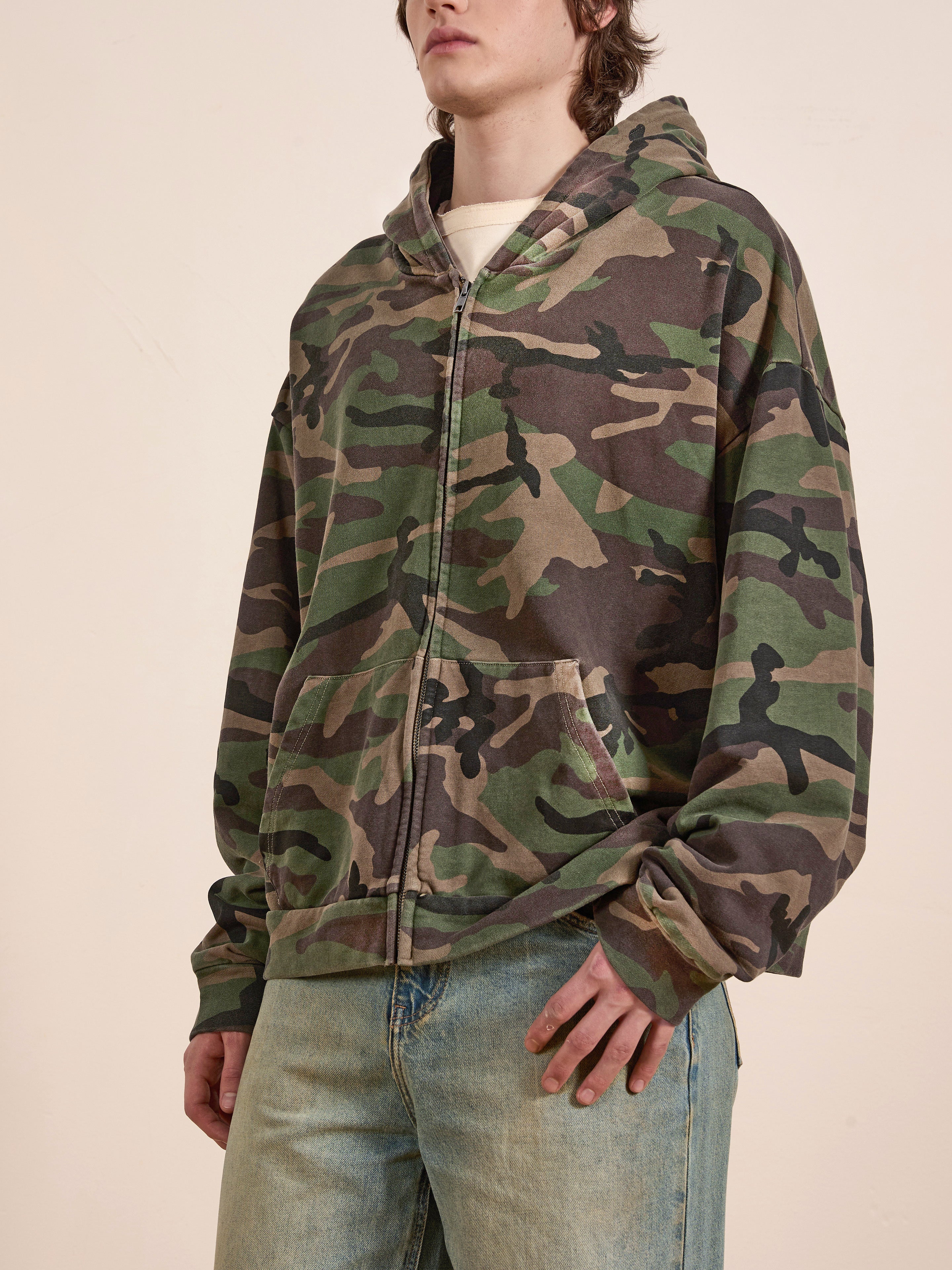 A person in a Found Washed Camouflage Zip Hoodie and blue jeans stands against a plain backdrop, the camo print adding a subtle edge to their look.