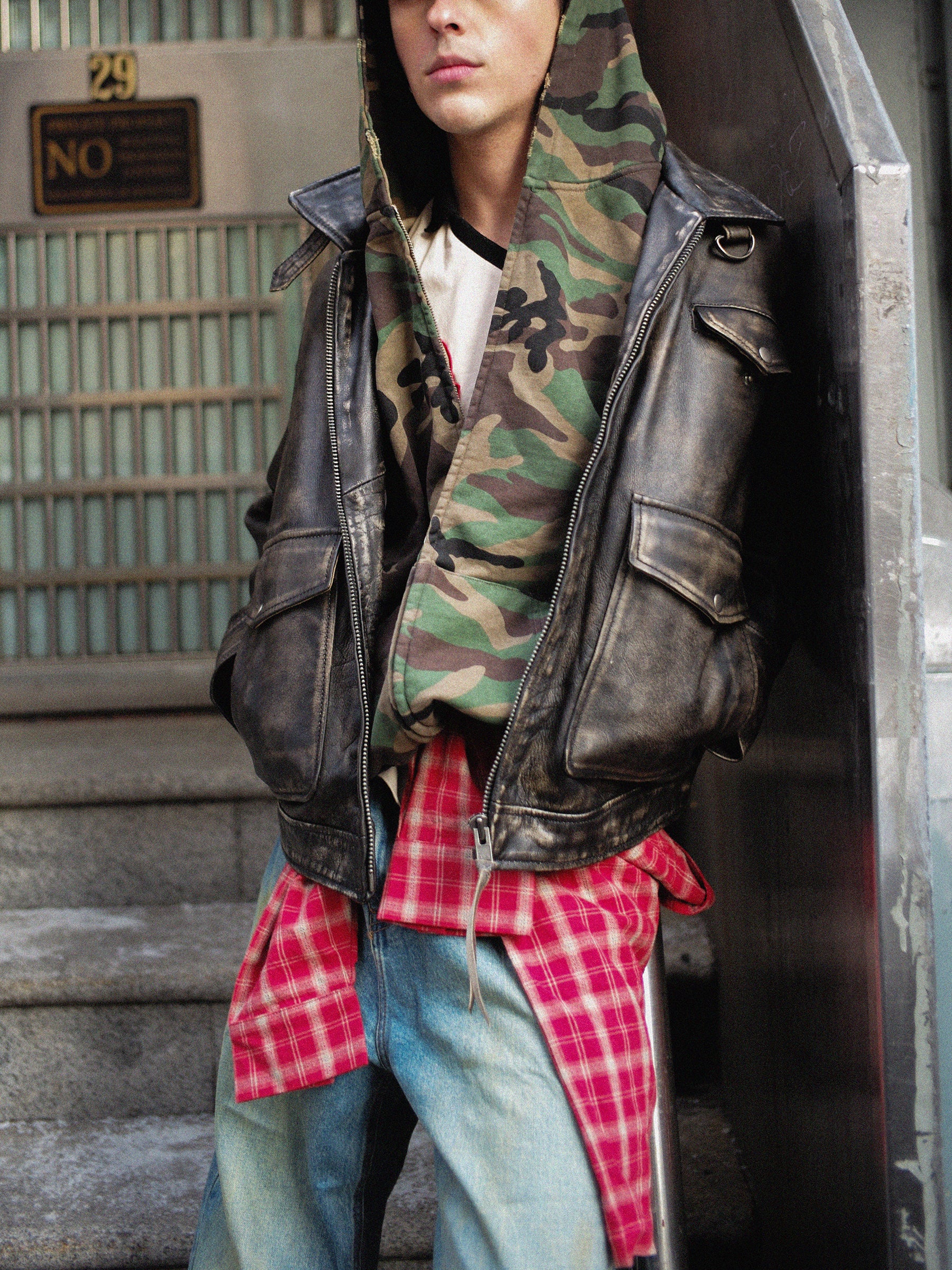 A person in a FOUND Washed Camouflage Zip Hoodie, plaid shirt, and leather jacket stands on steps.