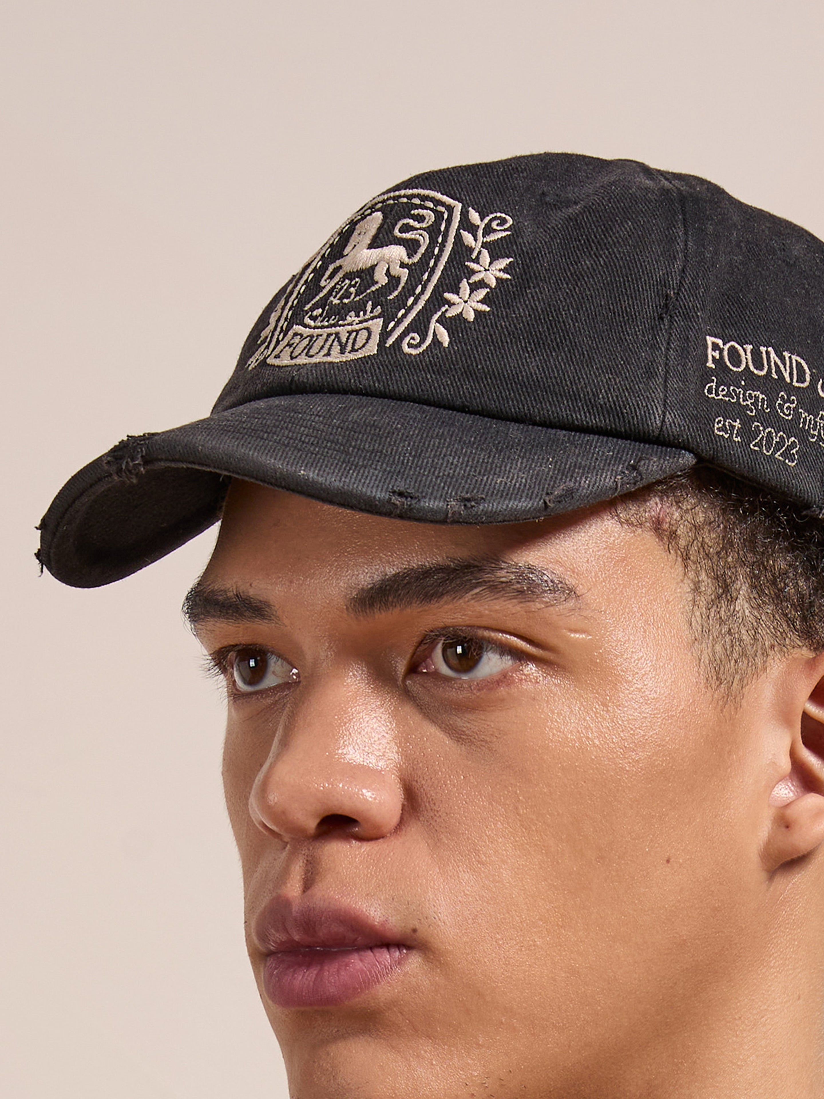 Someone wearing the FOUND Logo Crest Cap in washed black, featuring embroidered designs like a horse logo and the words "FOUND design & mgmt. est. 2023.
