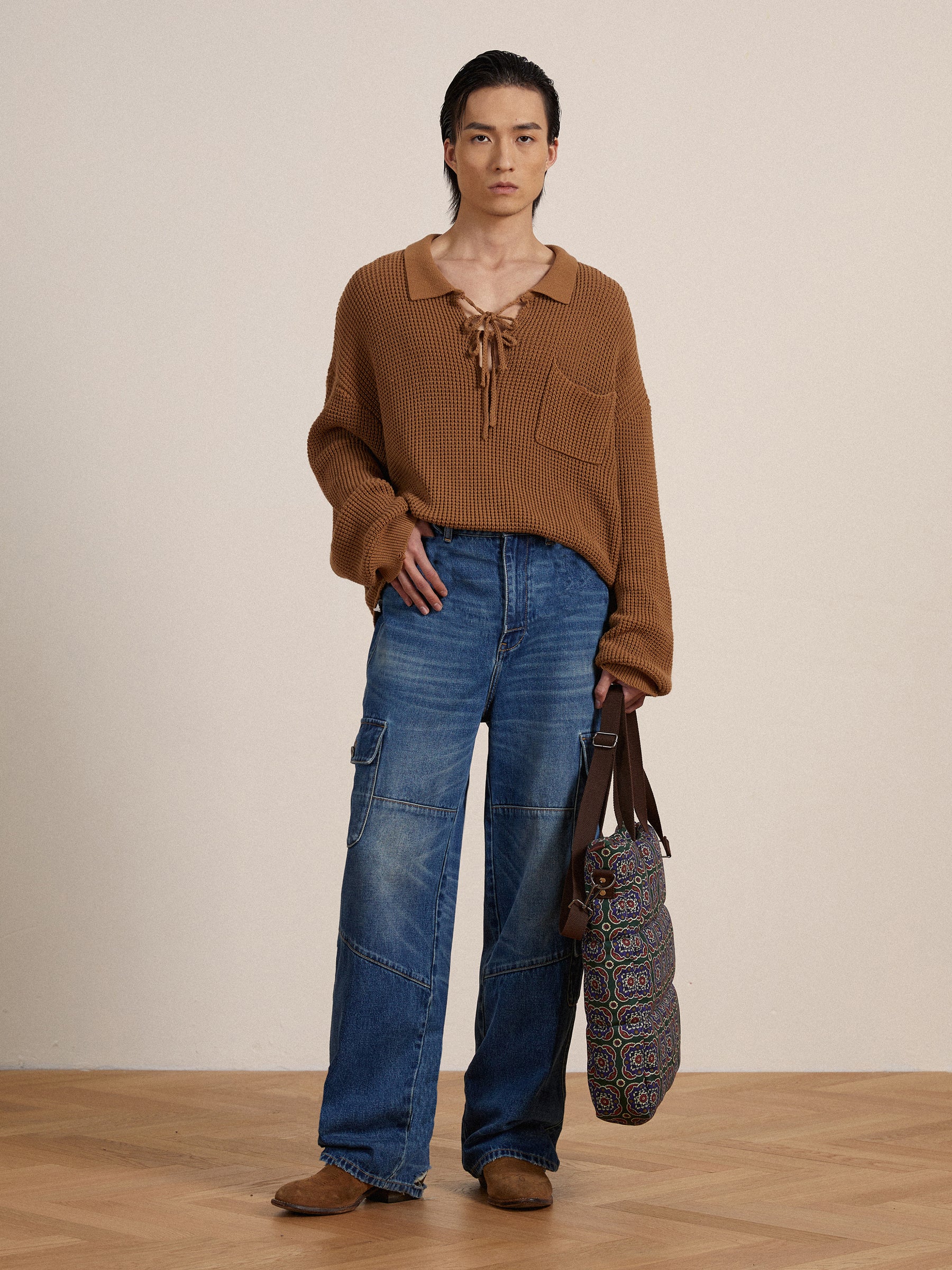 A person in a brown knitted top and Found Siwa Cargo Paneled Jeans, holding a patterned bag, stands in a neutral-toned room.