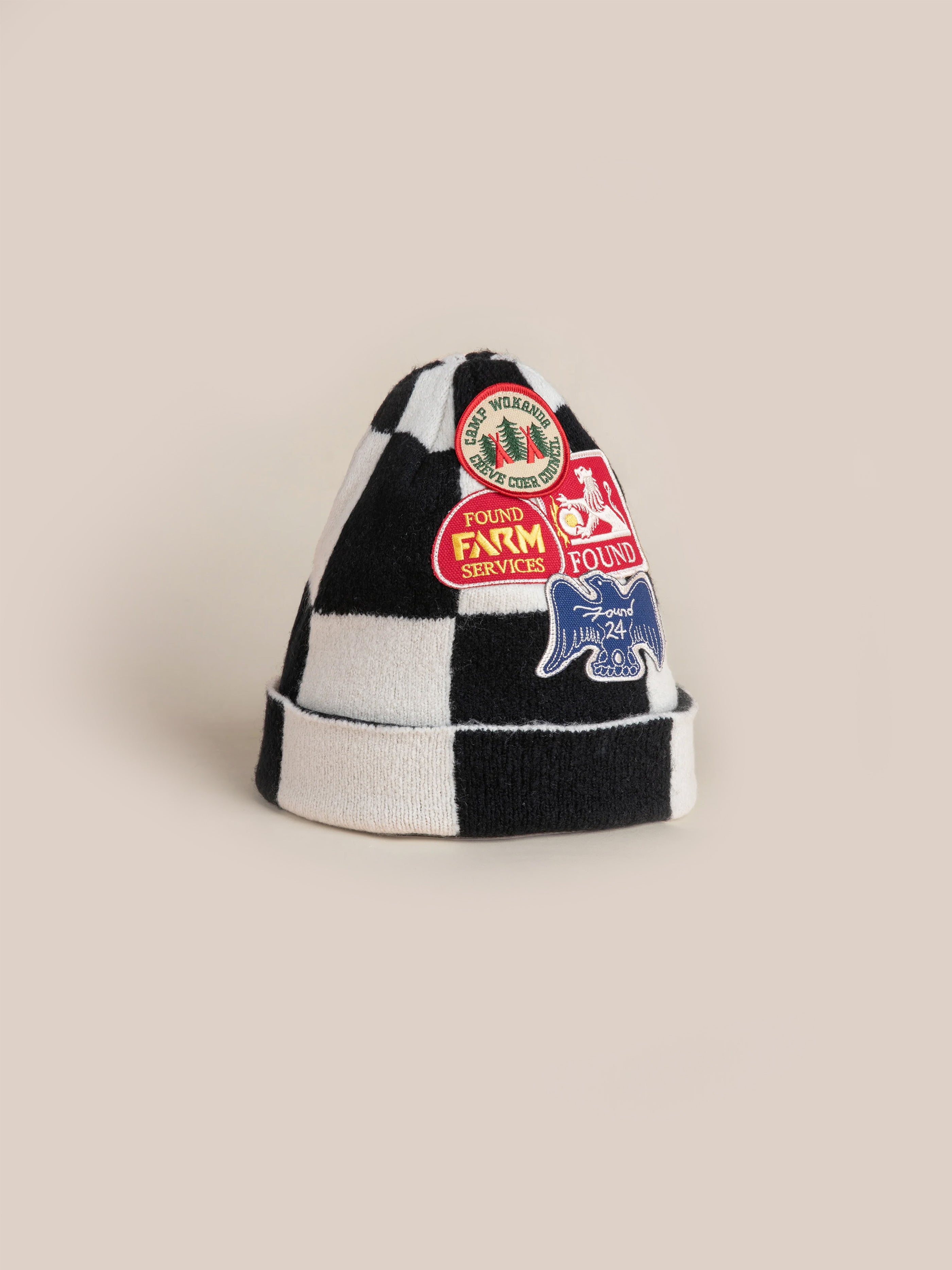 The Check Multi Patch Beanie by FOUND features a black and white checkered design crafted from a cozy knit blend, embellished with an assortment of colorful embroidered patches on a beige background.