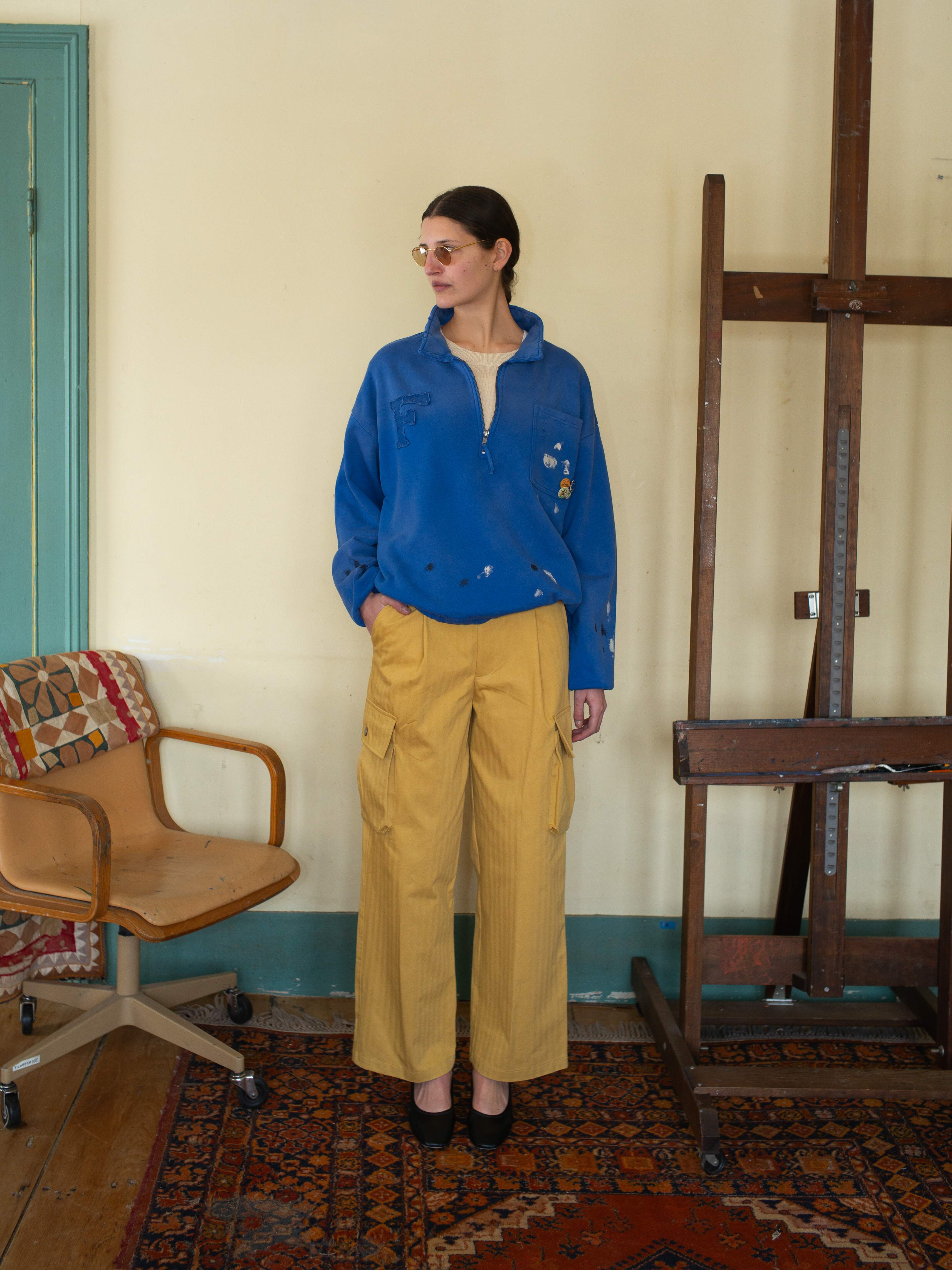 A person wearing a blue sweater and FOUND's Sunlight Twill Cargo Pants stands on a patterned rug near an easel and chair, with a beige wall in the background.